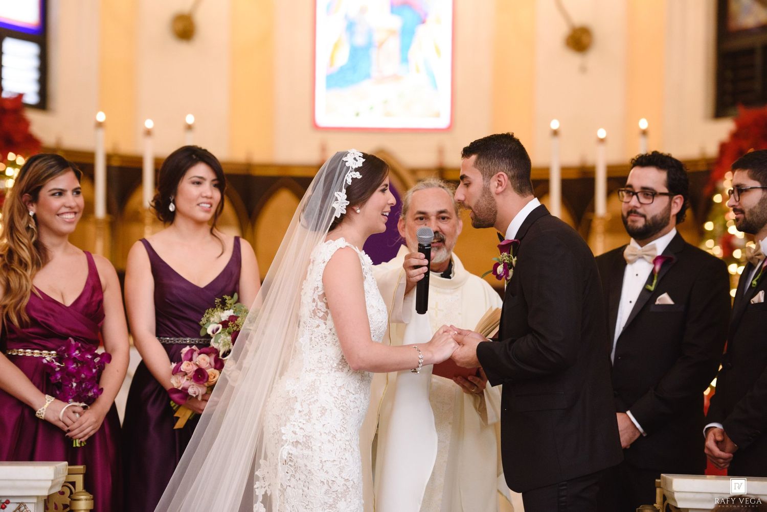 Bride and groom exchanging vows at church altar with bridesmaids in burgundy dresses and groomsmen during wedding ceremony.