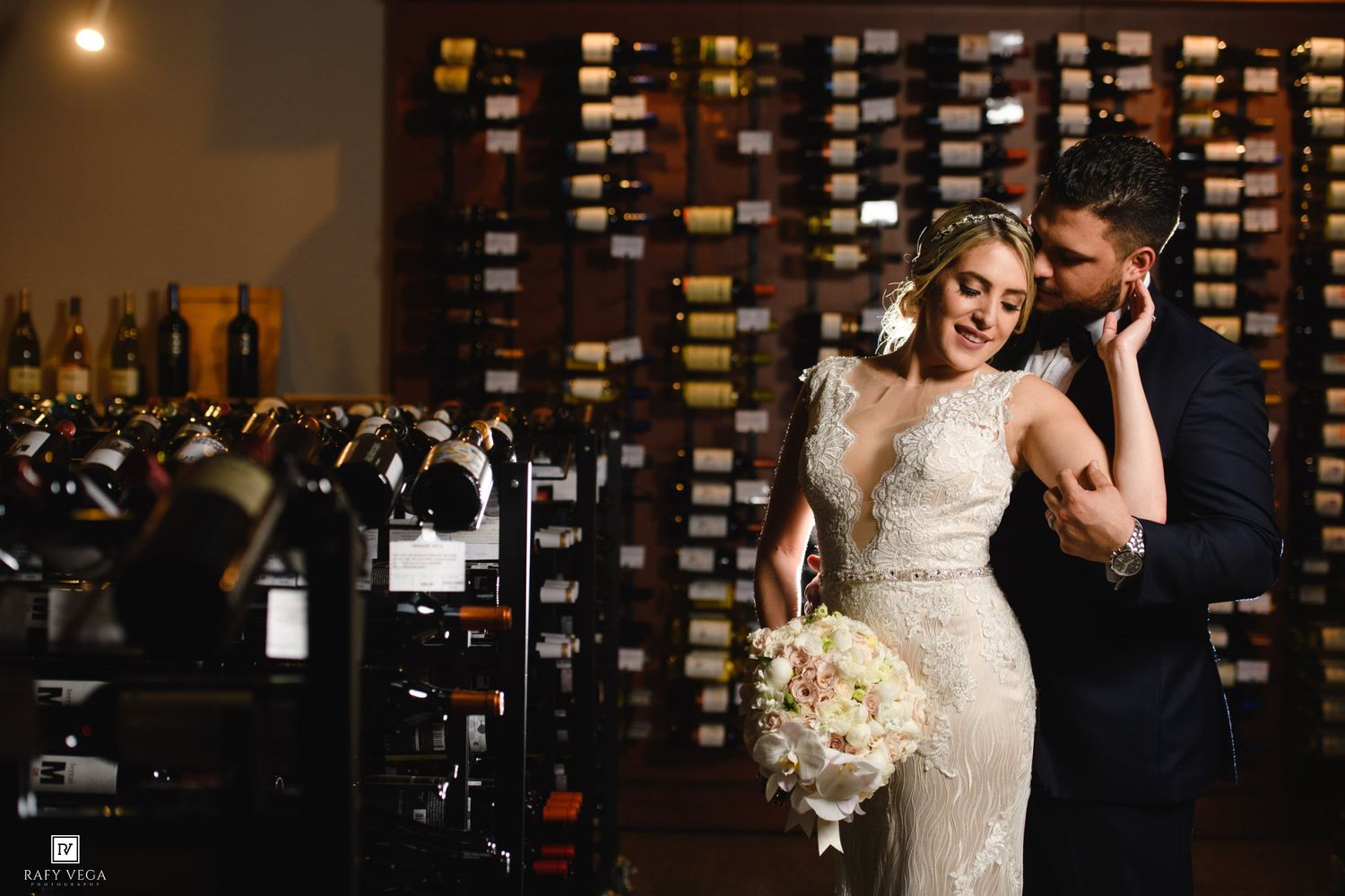 Bride and groom share a romantic moment in a wine cellar, surrounded by rows of wine bottles.