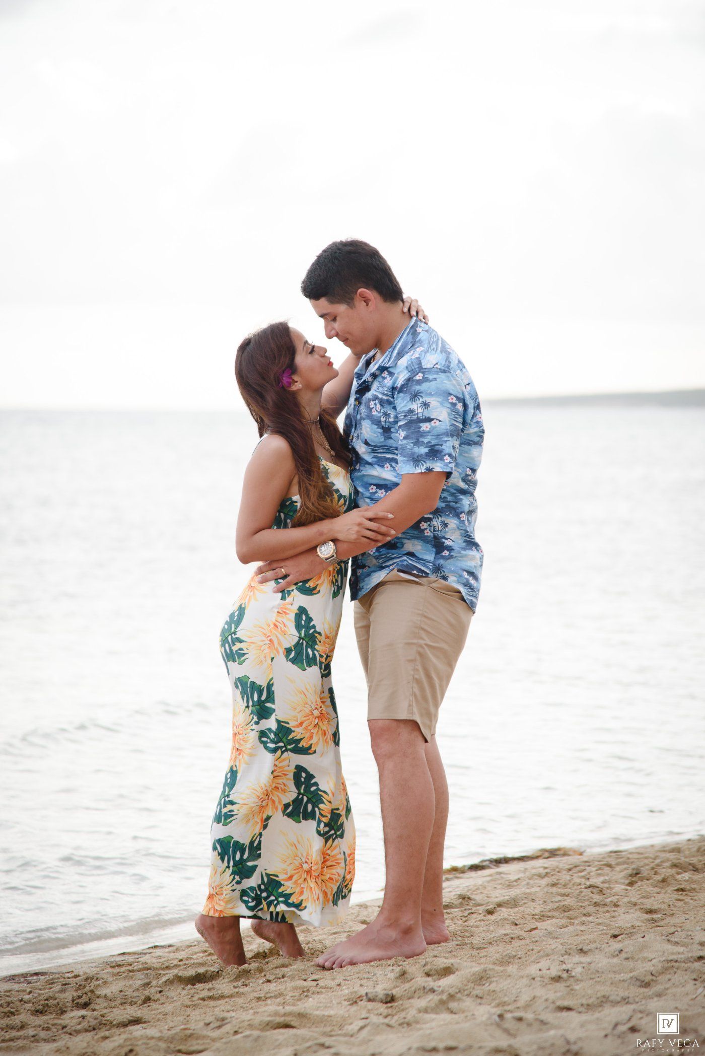 Couple embracing on a tropical beach, wearing floral outfits, sharing a romantic moment by the ocean shore.