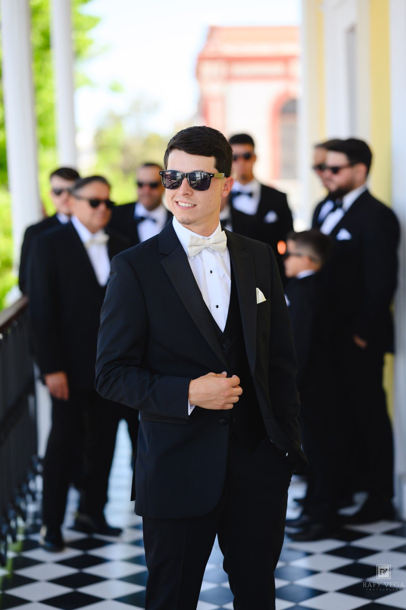 Groom in black tuxedo with white bow tie and sunglasses stands with groomsmen on checkered floor at wedding.
