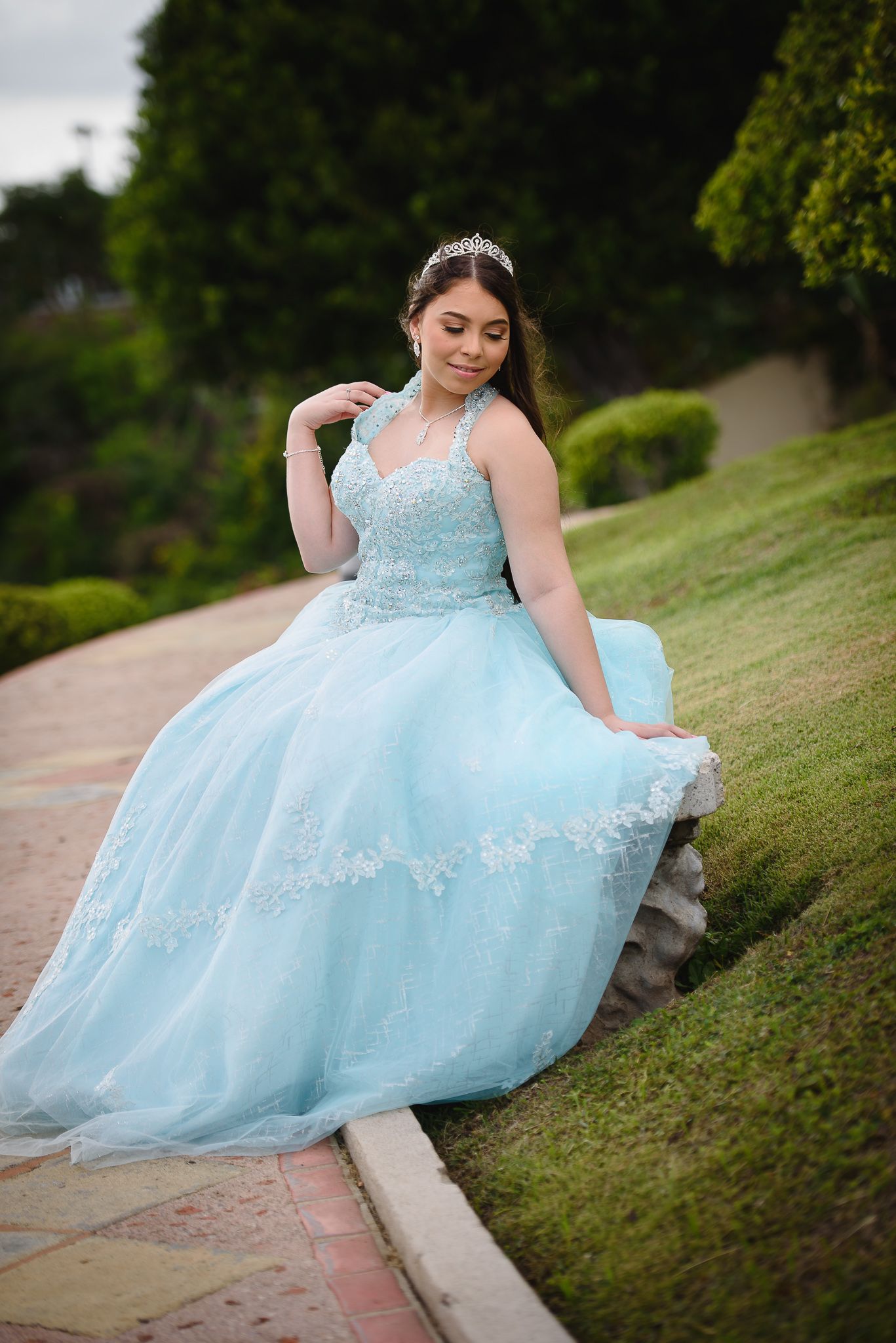 Young woman in a light blue ball gown with lace details and tiara poses gracefully in a garden setting.