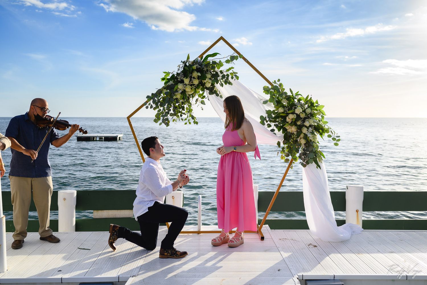 Man proposes to woman in pink dress on waterfront deck with floral arch and violinist at sunset.