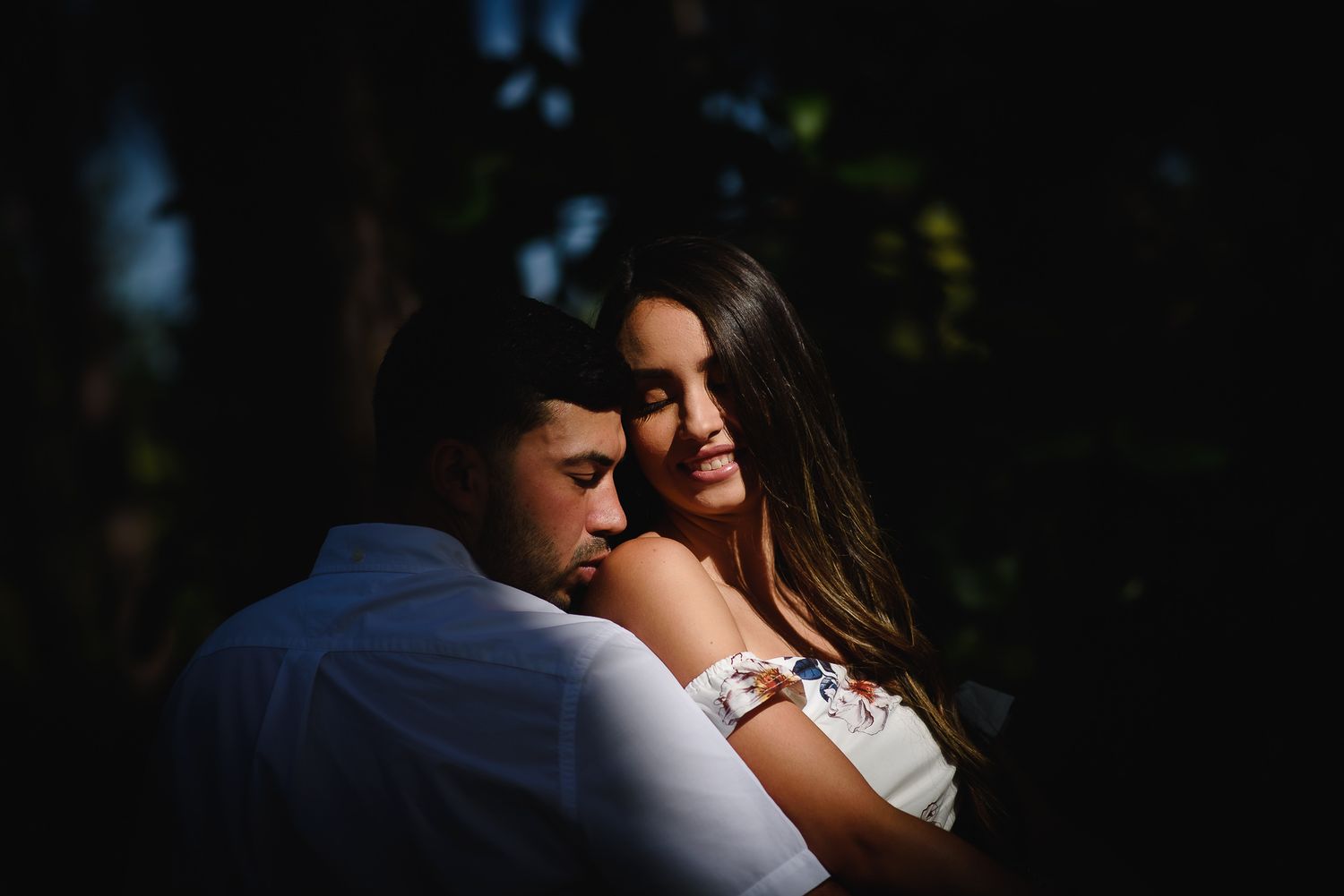 A couple embraces tenderly in soft sunlight, the woman smiling warmly as they share an intimate romantic moment outdoors.