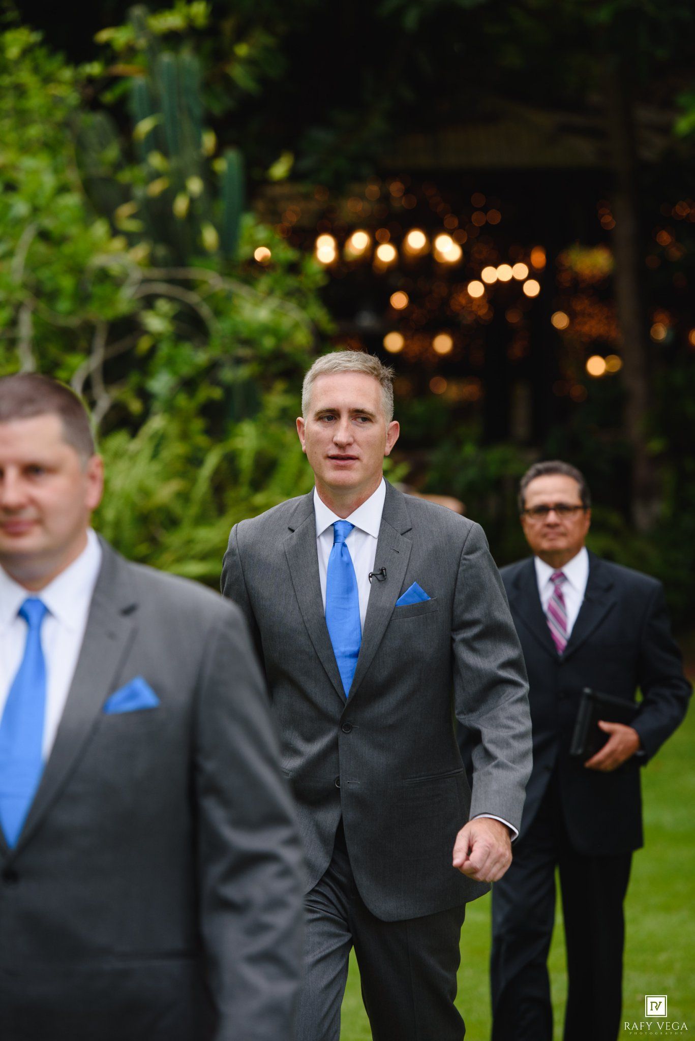 Three groomsmen in gray suits with blue ties walk through an outdoor wedding venue.