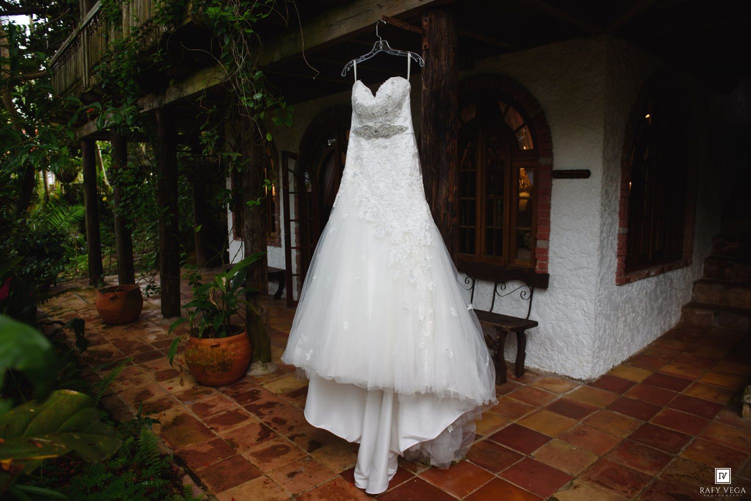 White strapless wedding gown with lace bodice hanging on display at a rustic outdoor venue with terracotta tiles.