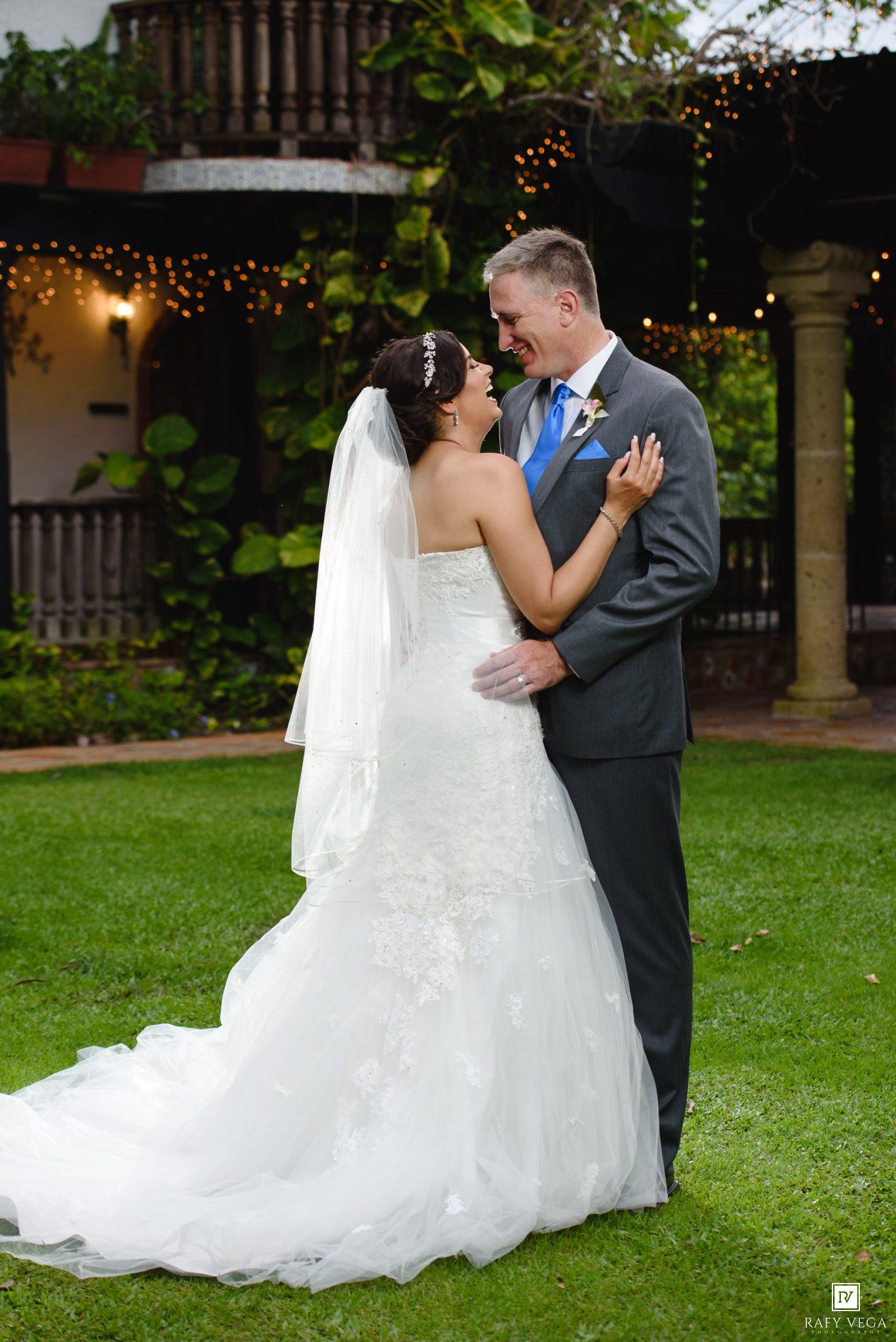 Bride and groom share a romantic moment on green lawn, she in lace gown with veil, he in gray suit with blue tie.