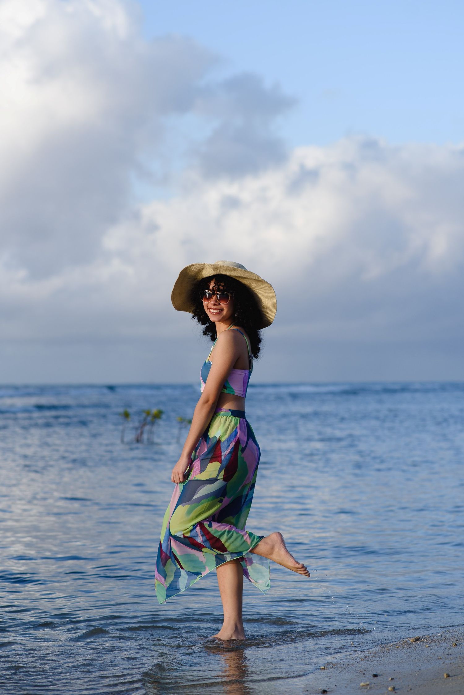 Woman in colorful dress, wide-brim hat, and sunglasses wading in ocean waves on a tropical beach.