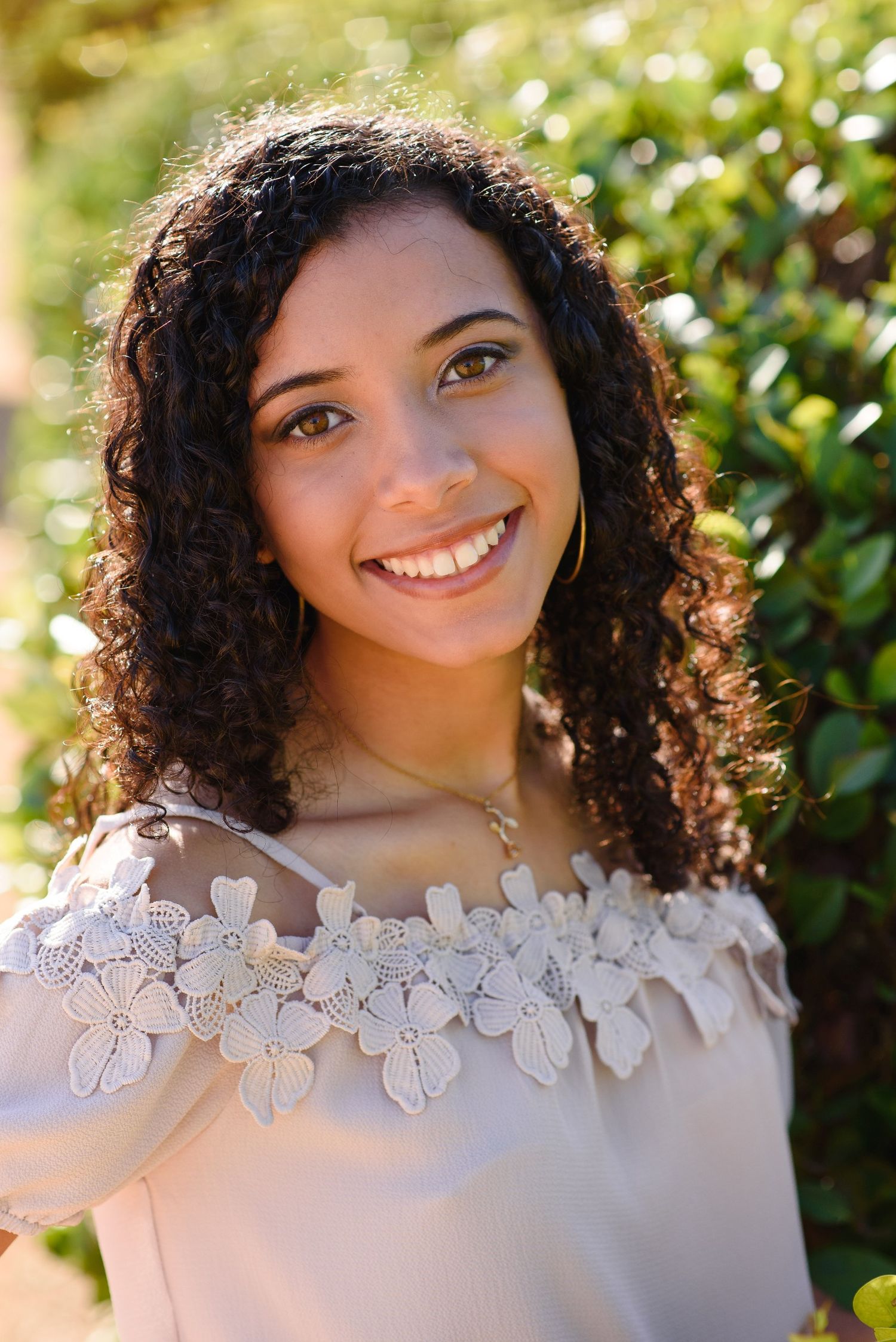Young woman with curly hair smiling outdoors, wearing a white floral off-shoulder top with green foliage background.
