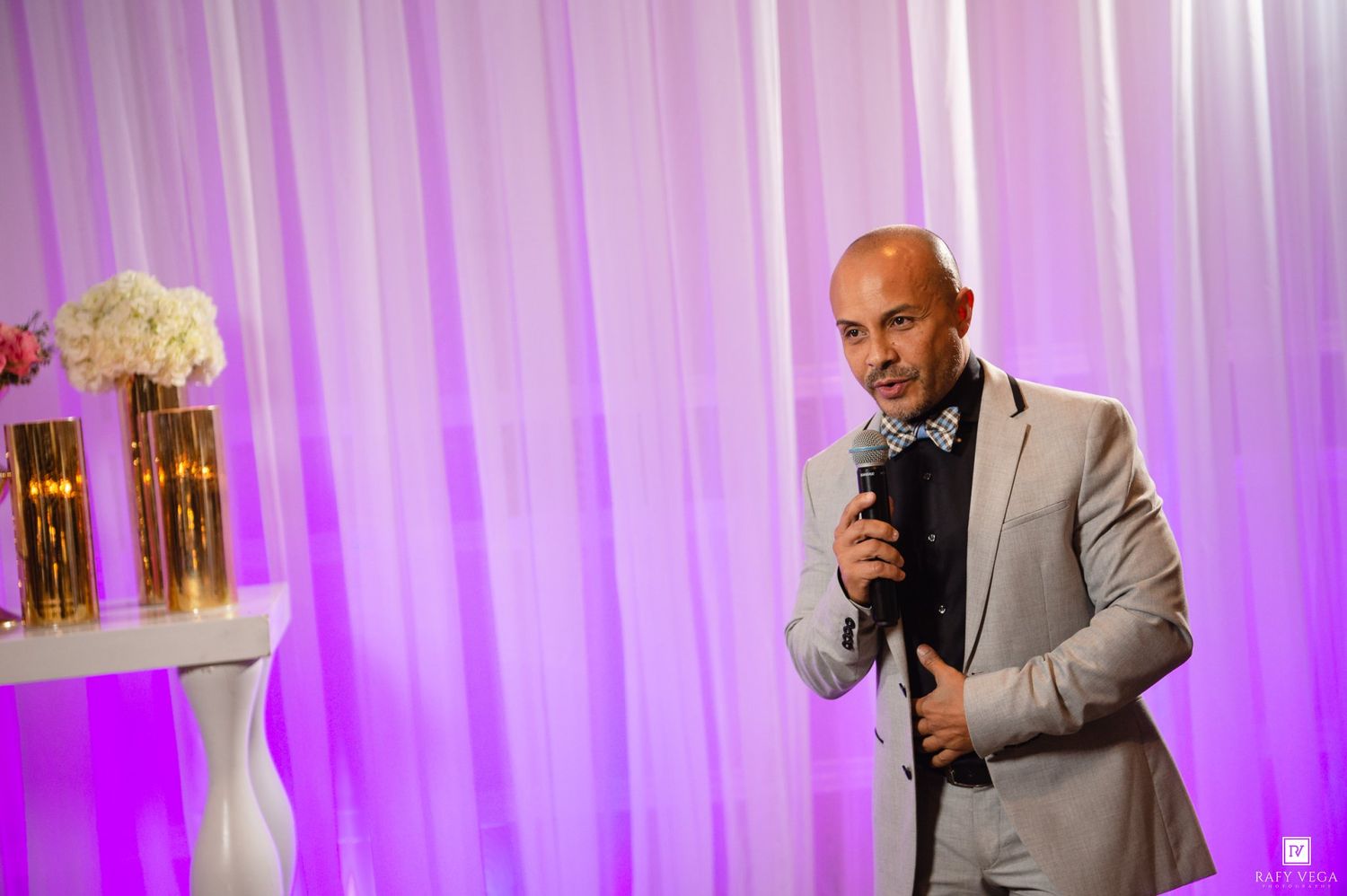 Bald man in gray suit speaking into microphone at elegant event with purple lighting and floral arrangements.