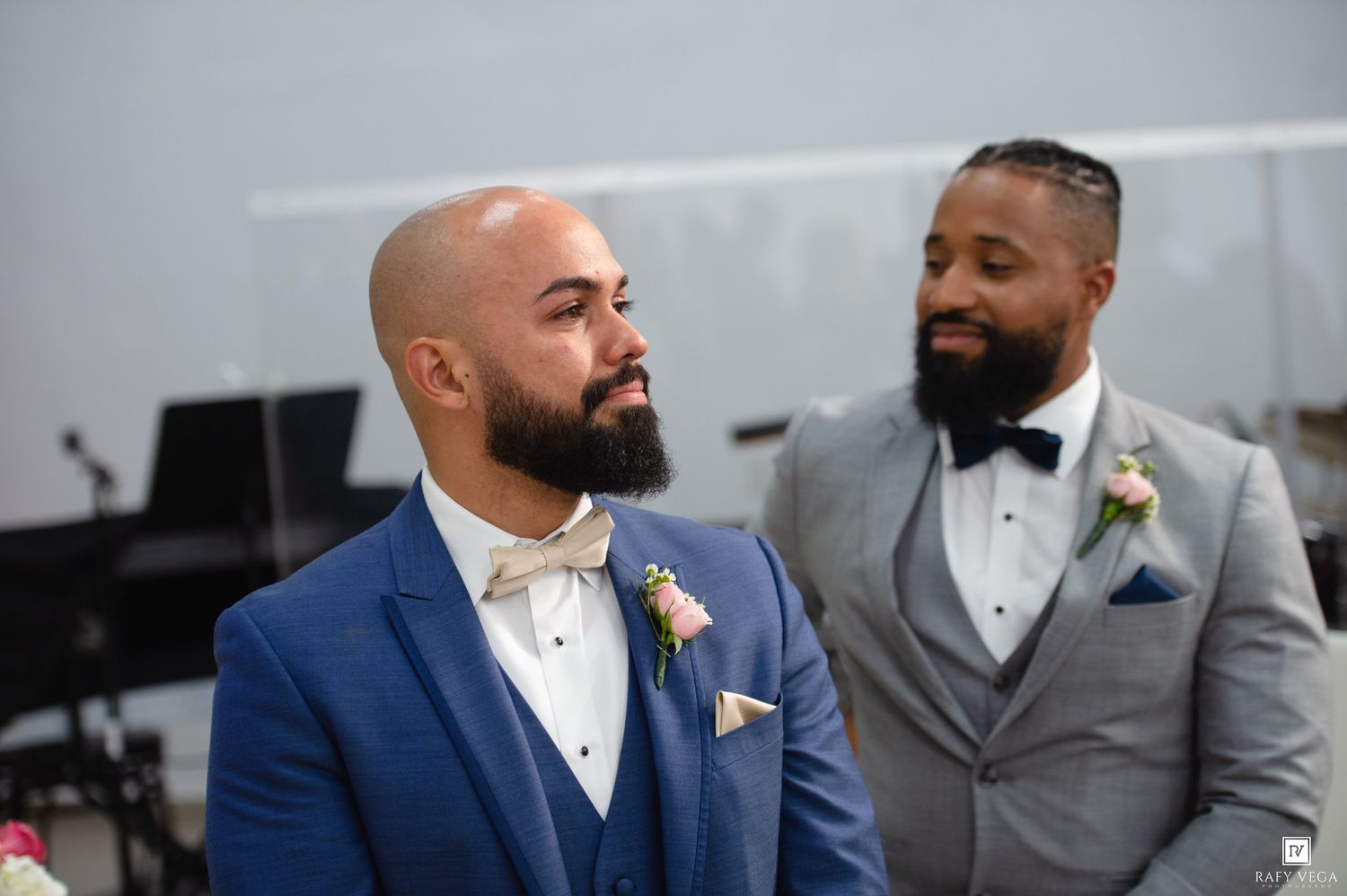 Two grooms in blue and gray suits with boutonnieres share a heartfelt moment during their wedding ceremony.