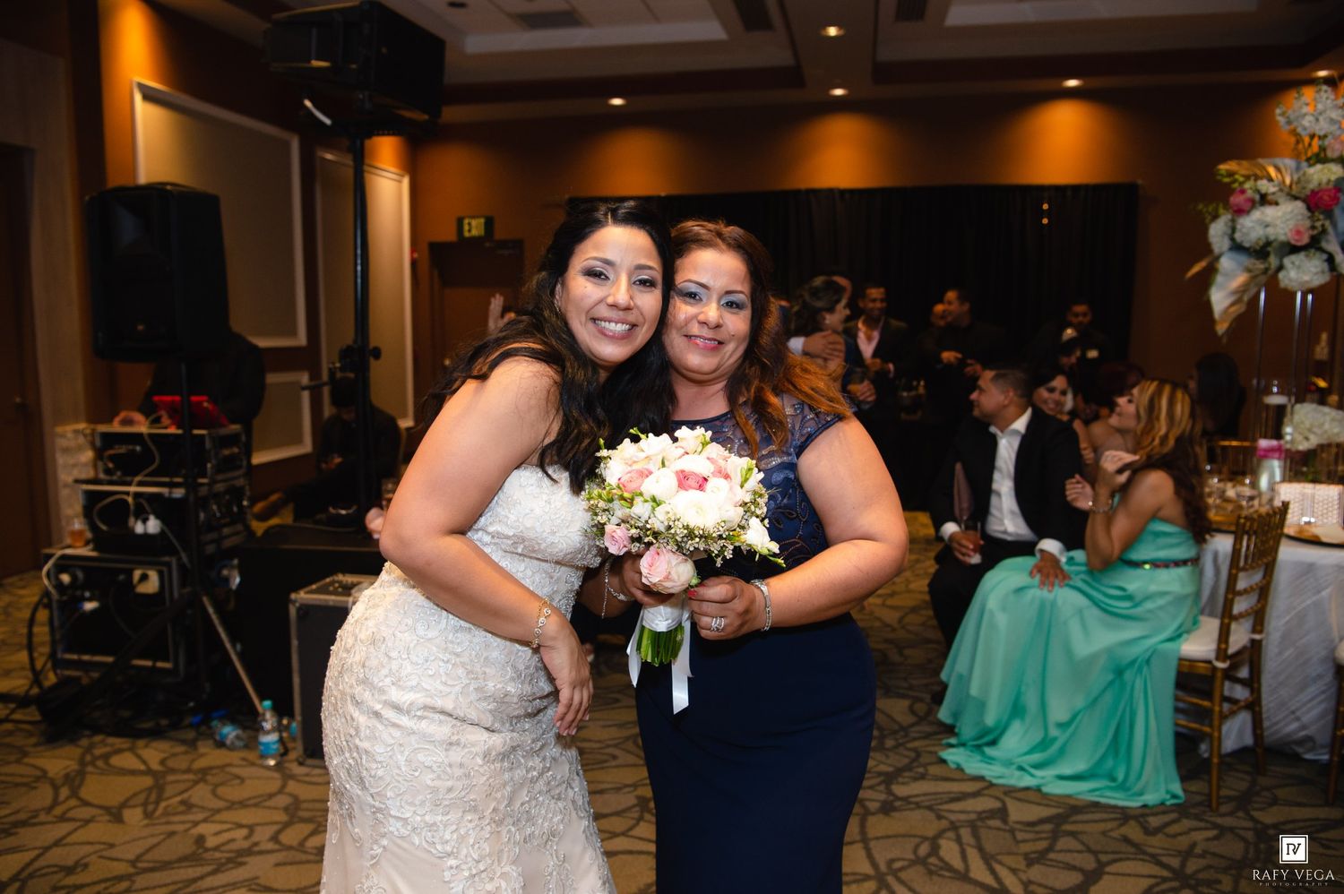 Bride in white lace gown and woman in navy dress smile together holding bouquet at wedding reception.