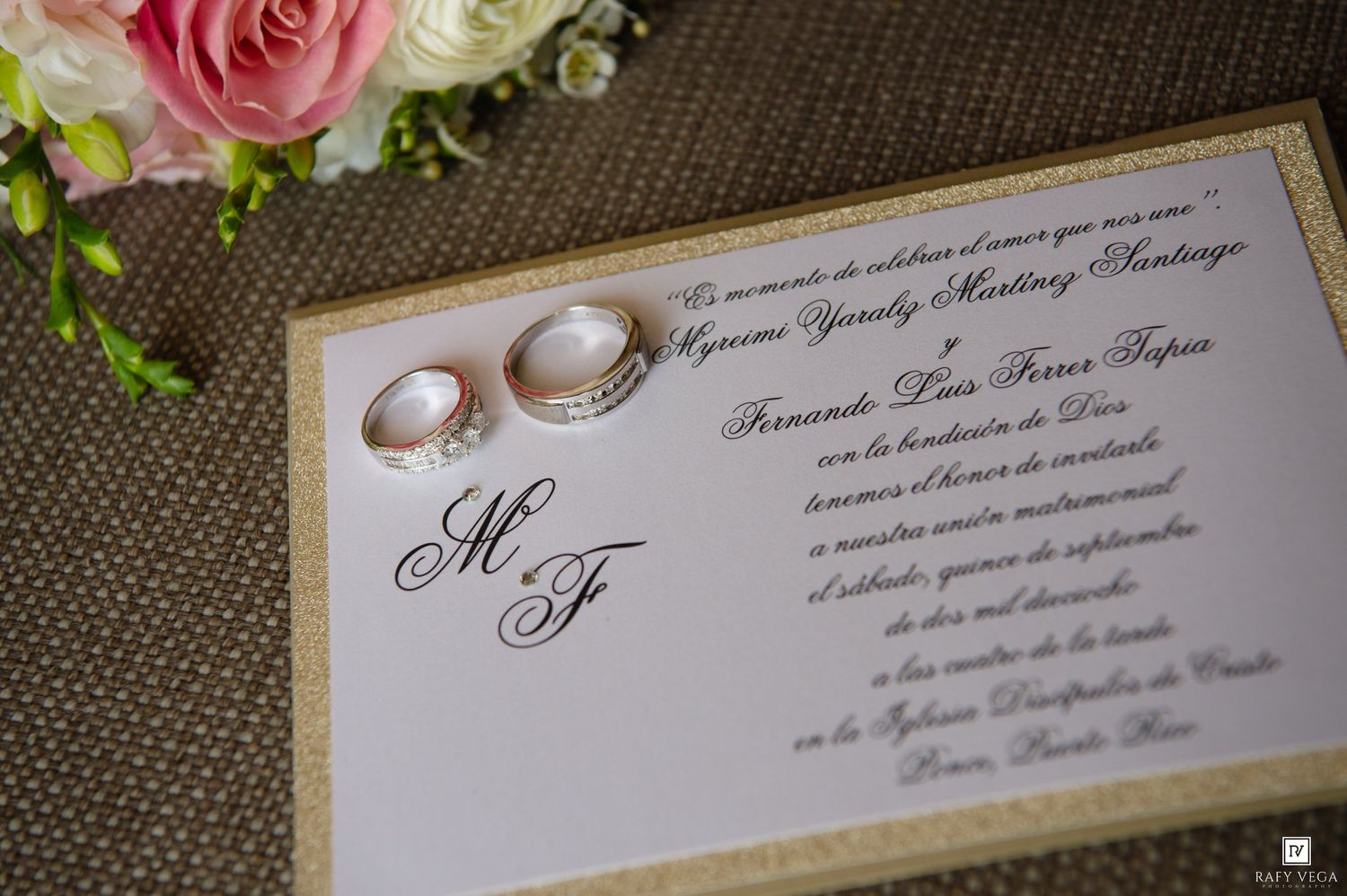 Elegant wedding invitation with gold border, monogram MF, and two pearl rings resting on top, with pink roses in background.