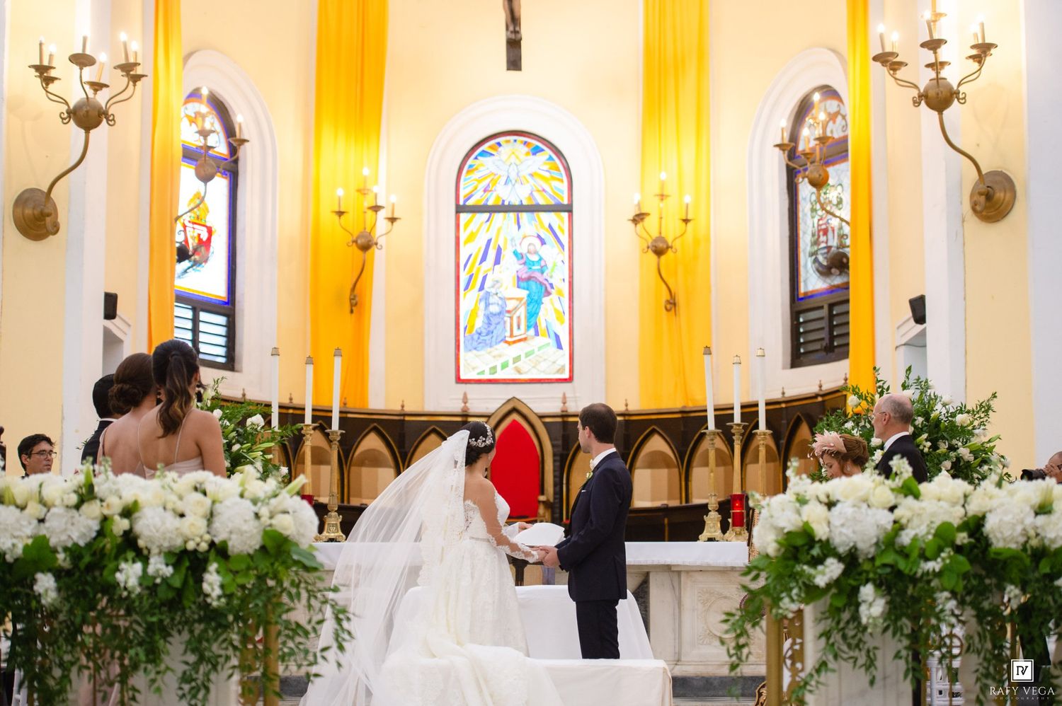 Bride and groom exchange vows at a church altar adorned with white floral arrangements and colorful stained glass windows.