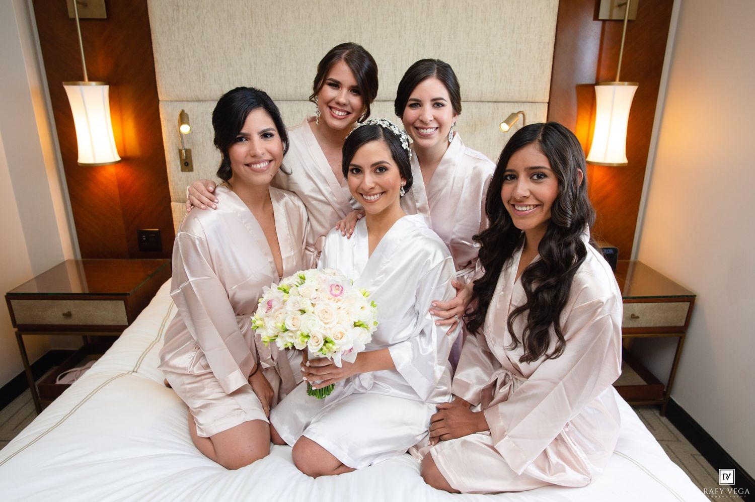 Bride in white robe holding bouquet surrounded by four bridesmaids in matching robes sitting on hotel bed.
