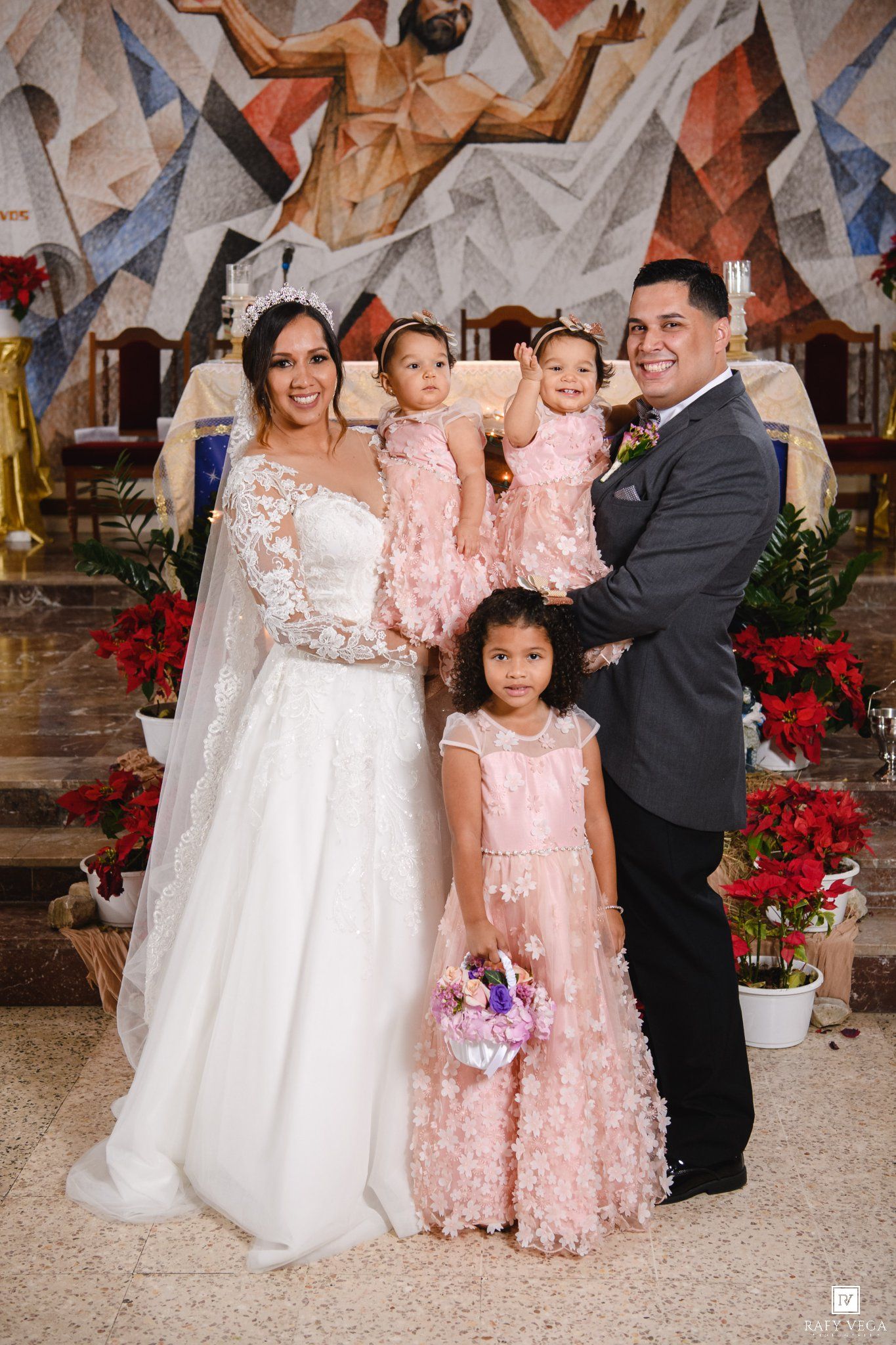 Wedding couple posing with three girls in pink dresses inside a decorated church altar.
