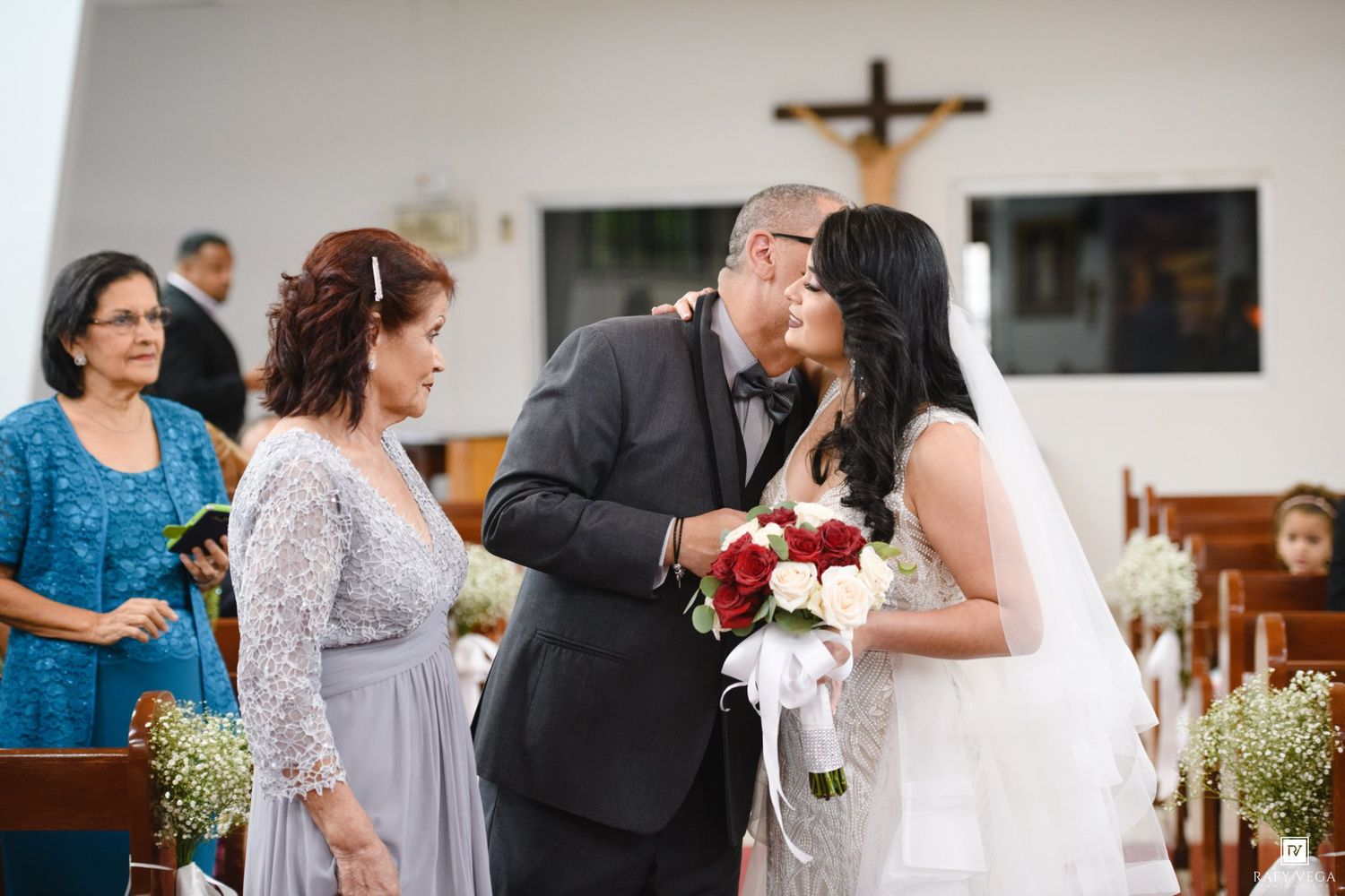 Father kissing bride at church altar during wedding ceremony with mother standing nearby.