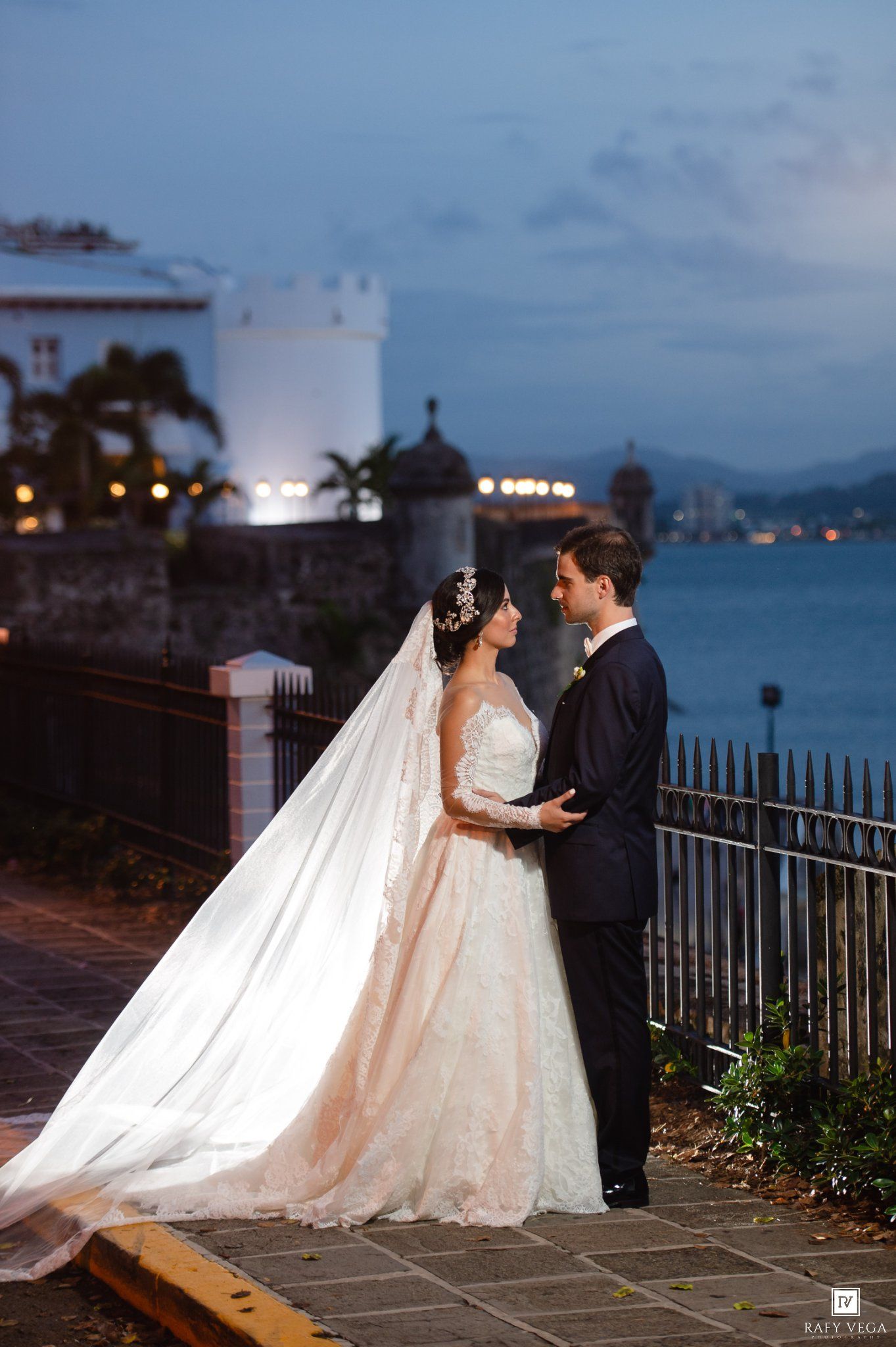 Bride and groom share a romantic moment at dusk by a waterfront railing, bride wearing a flowing lace gown and long veil.