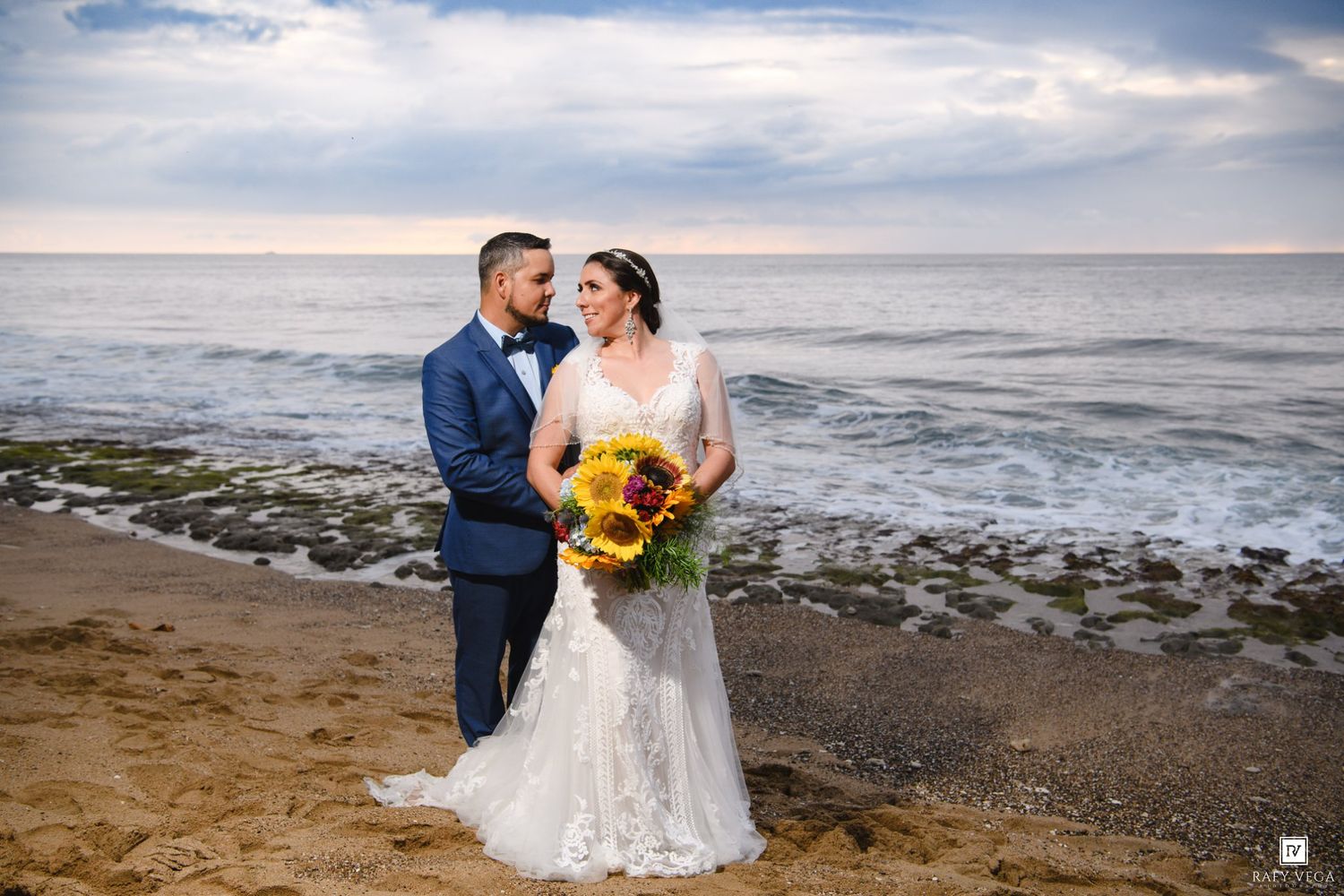 Bride and groom embrace on sandy beach at sunset, bride holding vibrant sunflower bouquet in lace wedding gown.