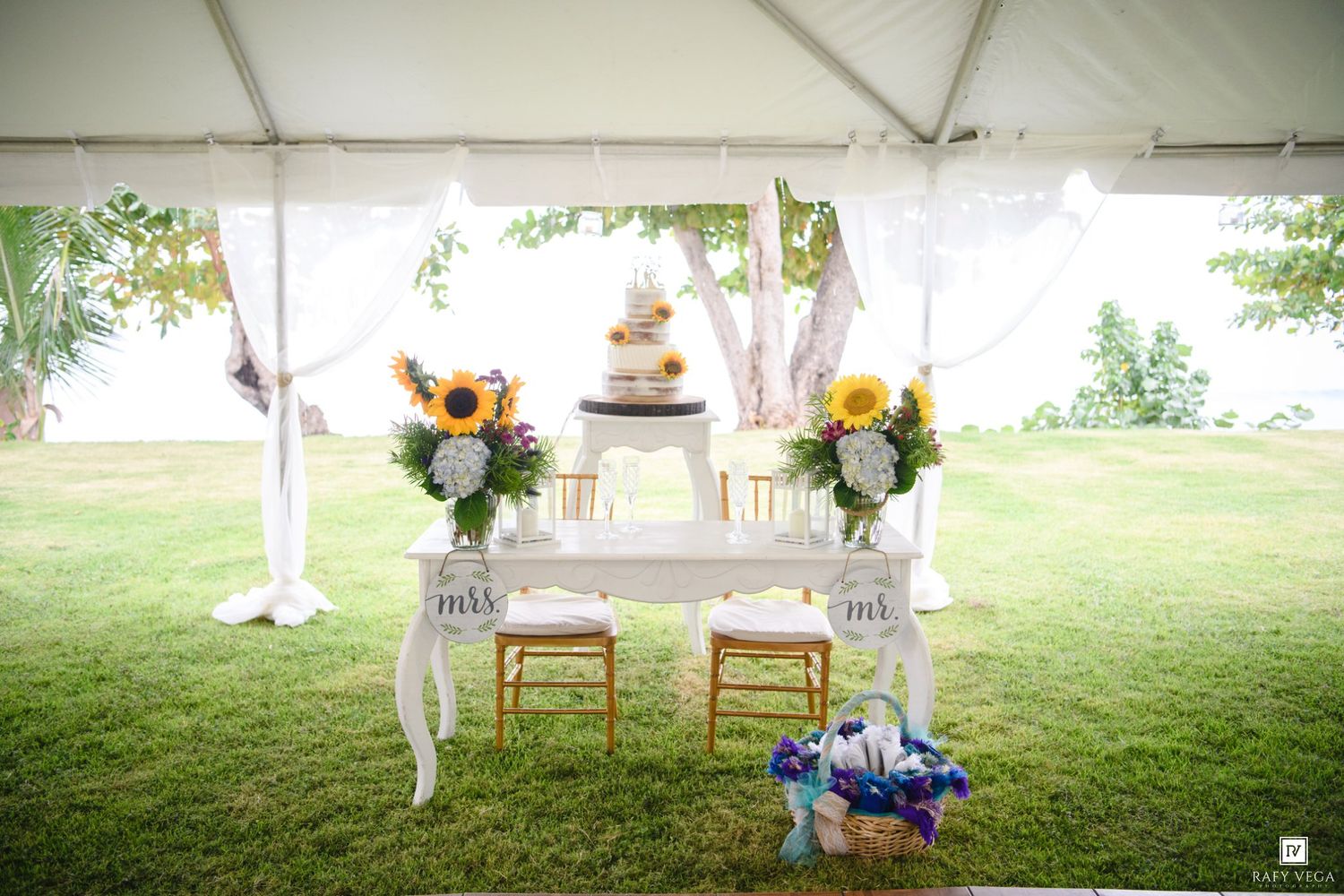 Elegant wedding sweetheart table with Mr. and Mrs. signs, sunflower arrangements, and tiered cake under a white tent.