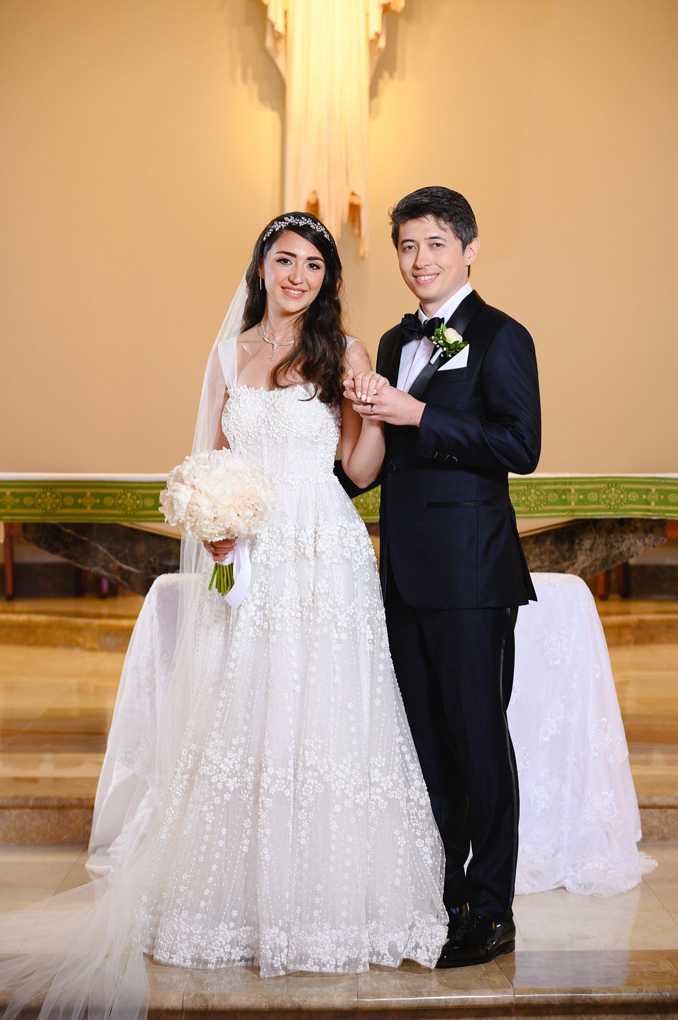 Bride in floral gown and groom in tuxedo hold hands while posing at a church altar.