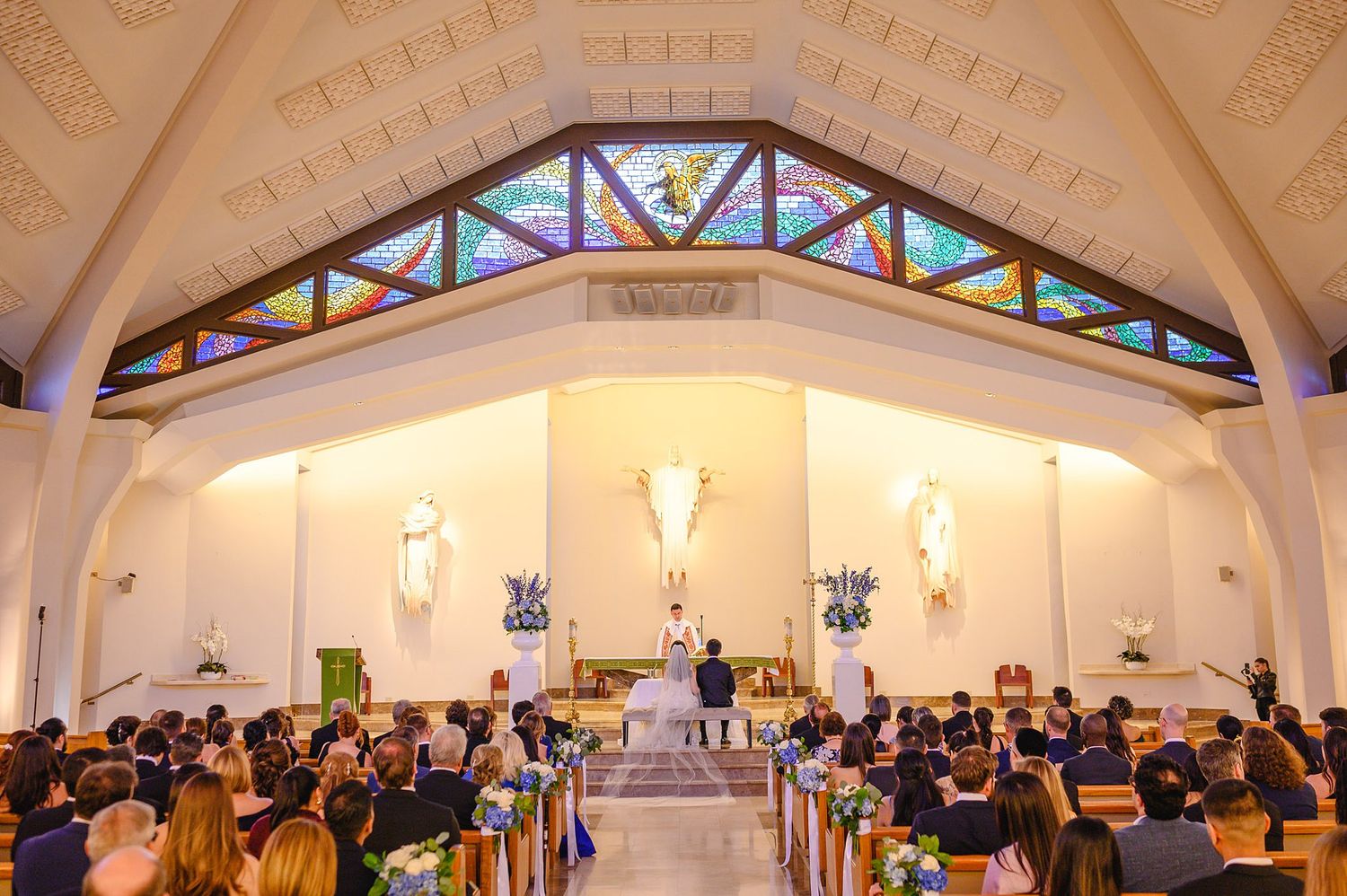 Wedding ceremony inside a beautiful church with stained glass windows, wooden pews filled with guests, and couple at the altar.