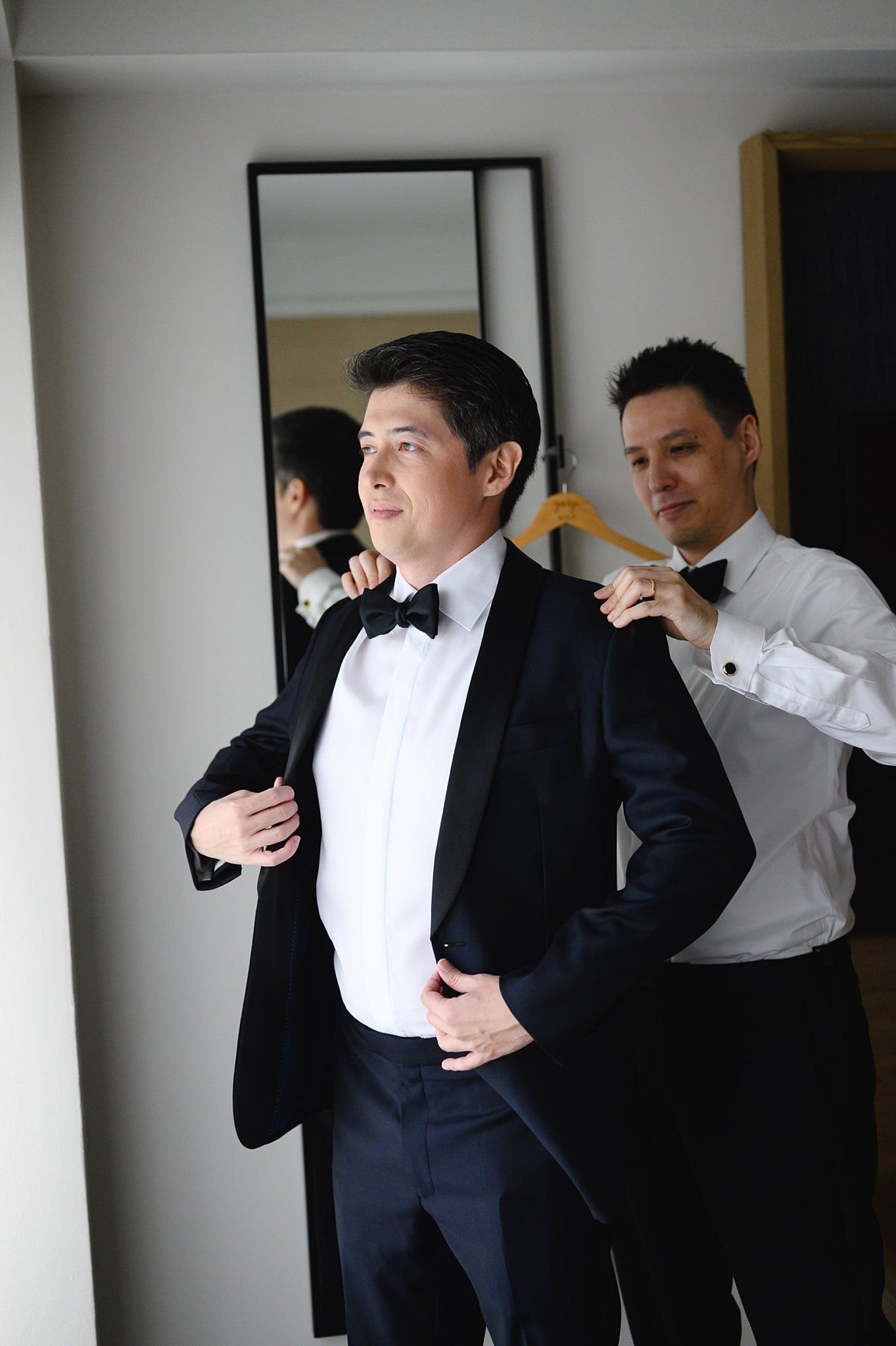 Groom in black tuxedo getting jacket adjusted by groomsman, both wearing bow ties, preparing for wedding ceremony.