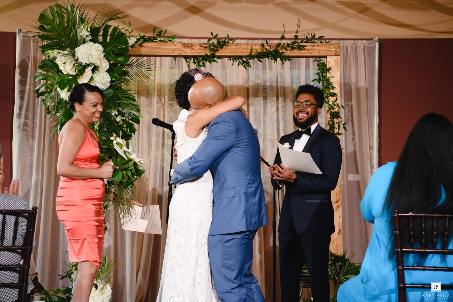 Bride and groom share first kiss at wedding altar decorated with greenery and white flowers, officiant smiling nearby.