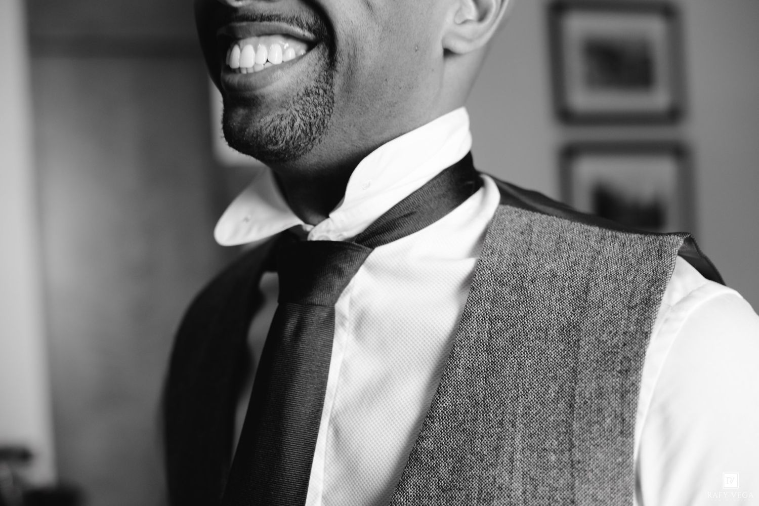 Black and white photo of a smiling groom adjusting his tie, wearing a tweed vest and white dress shirt.