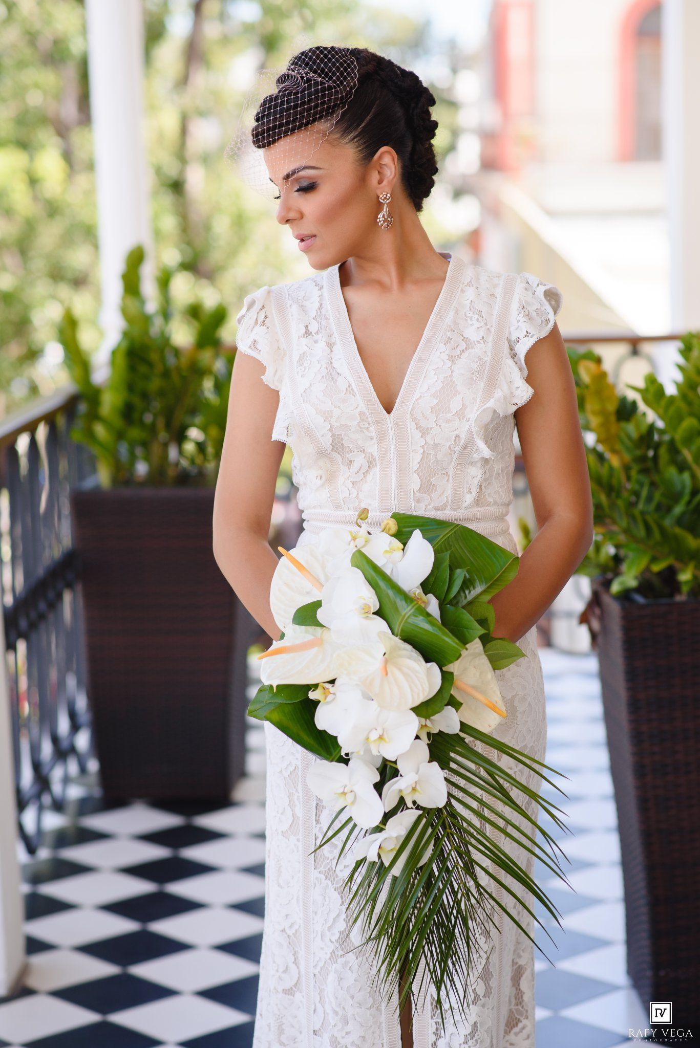 Elegant bride in white lace dress holding tropical bouquet with orchids on a checkered balcony.