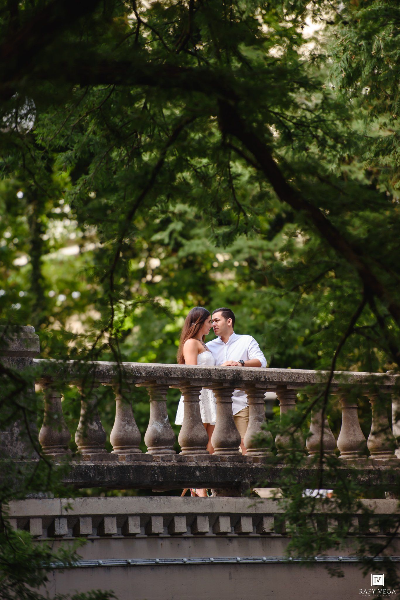 Couple sharing a romantic moment on a stone balustrade bridge surrounded by lush green trees.