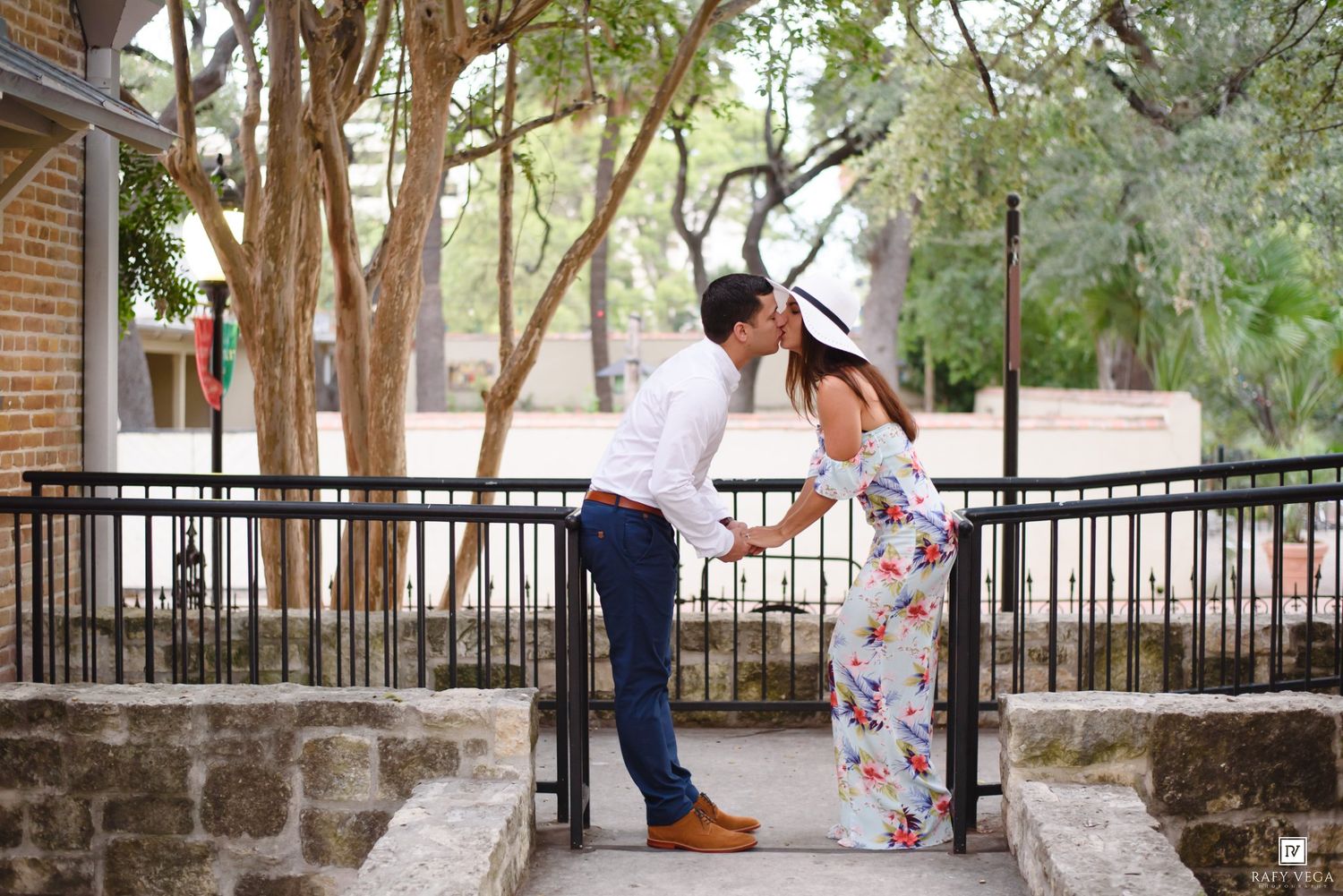Couple sharing a romantic kiss near iron railings in an outdoor park setting, woman in floral dress and sun hat.
