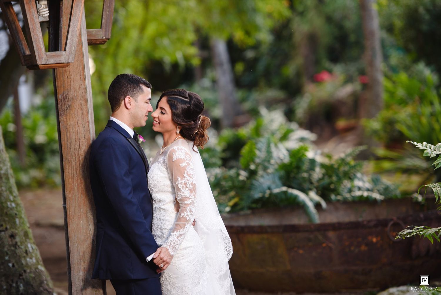 A bride and groom share a romantic kiss outdoors, surrounded by lush greenery, the bride in lace gown and groom in navy suit.