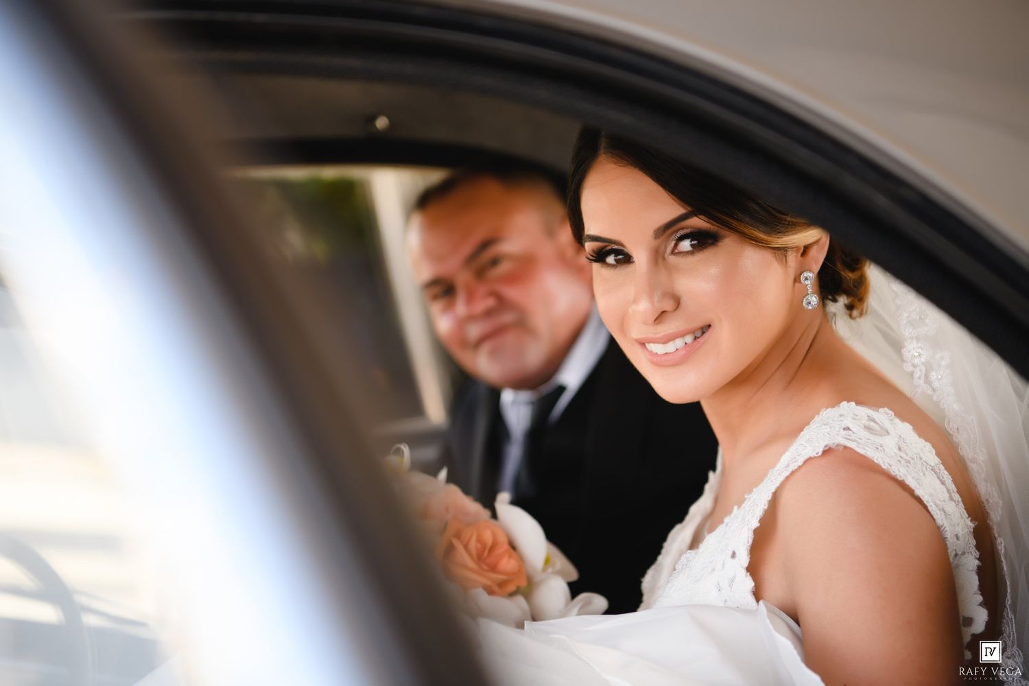 Bride and groom sitting in a car on their wedding day, bride smiling in white lace dress with pearl earrings.