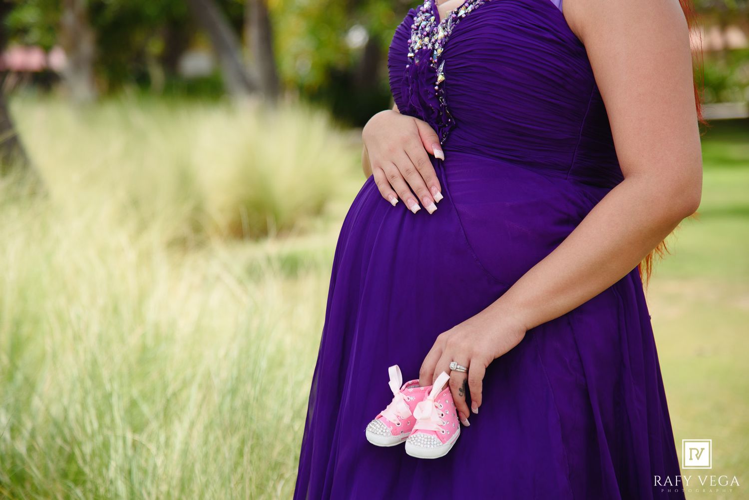 Pregnant woman in purple dress holding pink baby shoes outdoors during maternity photo shoot.