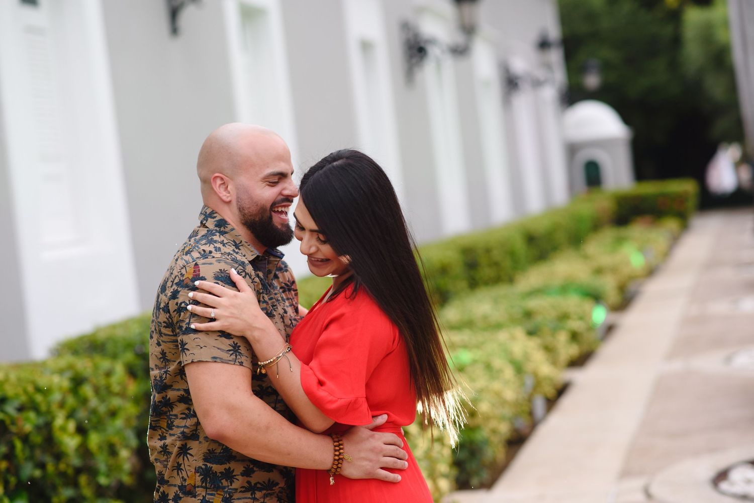 A couple embraces and smiles at each other outdoors, man in camo shirt and woman in red dress, lush green hedges behind them.