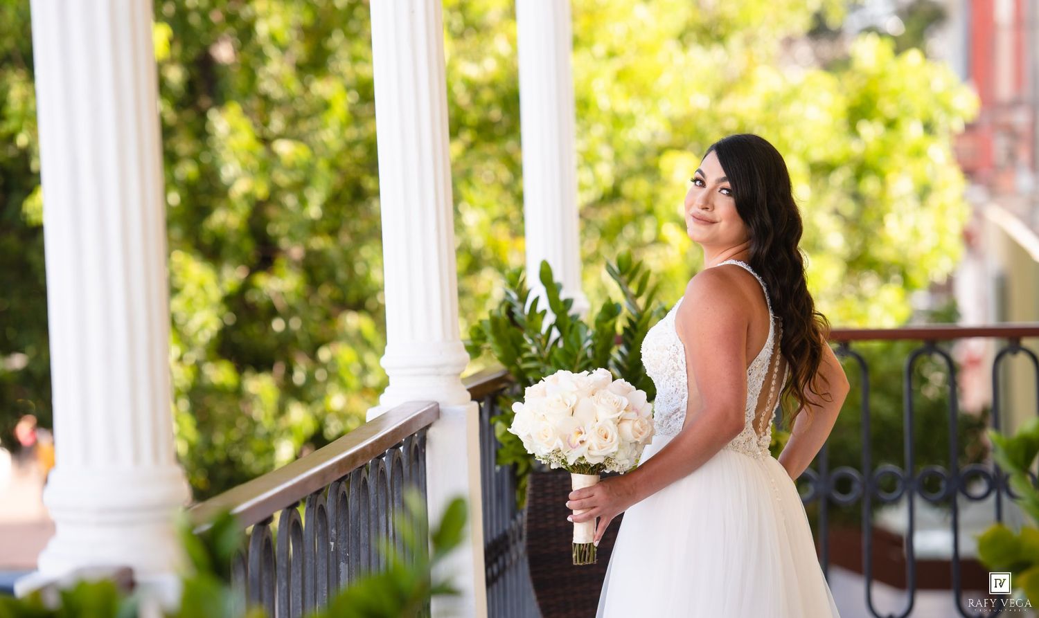 Bride in white gown holding white bouquet on elegant porch with columns and lush greenery.