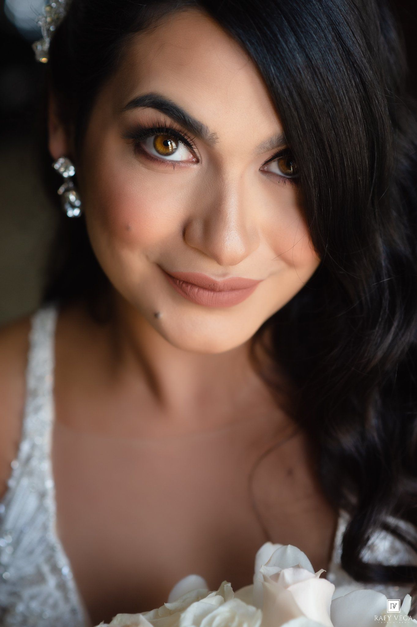 Close-up portrait of a beautiful bride with dark hair, amber eyes, diamond earrings, and natural glam makeup holding white roses.