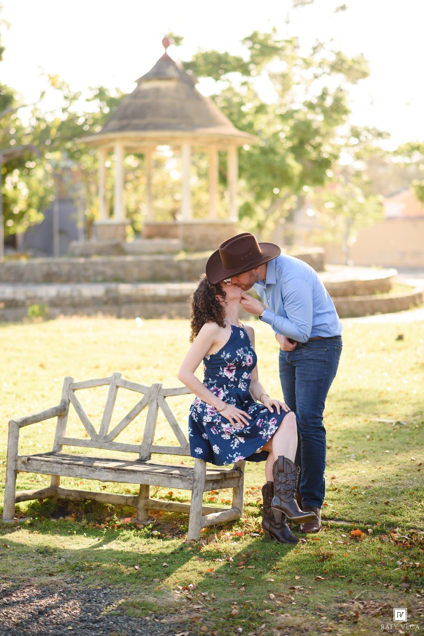 Couple sharing a romantic moment on a rustic bench in a sunlit park with a gazebo backdrop.