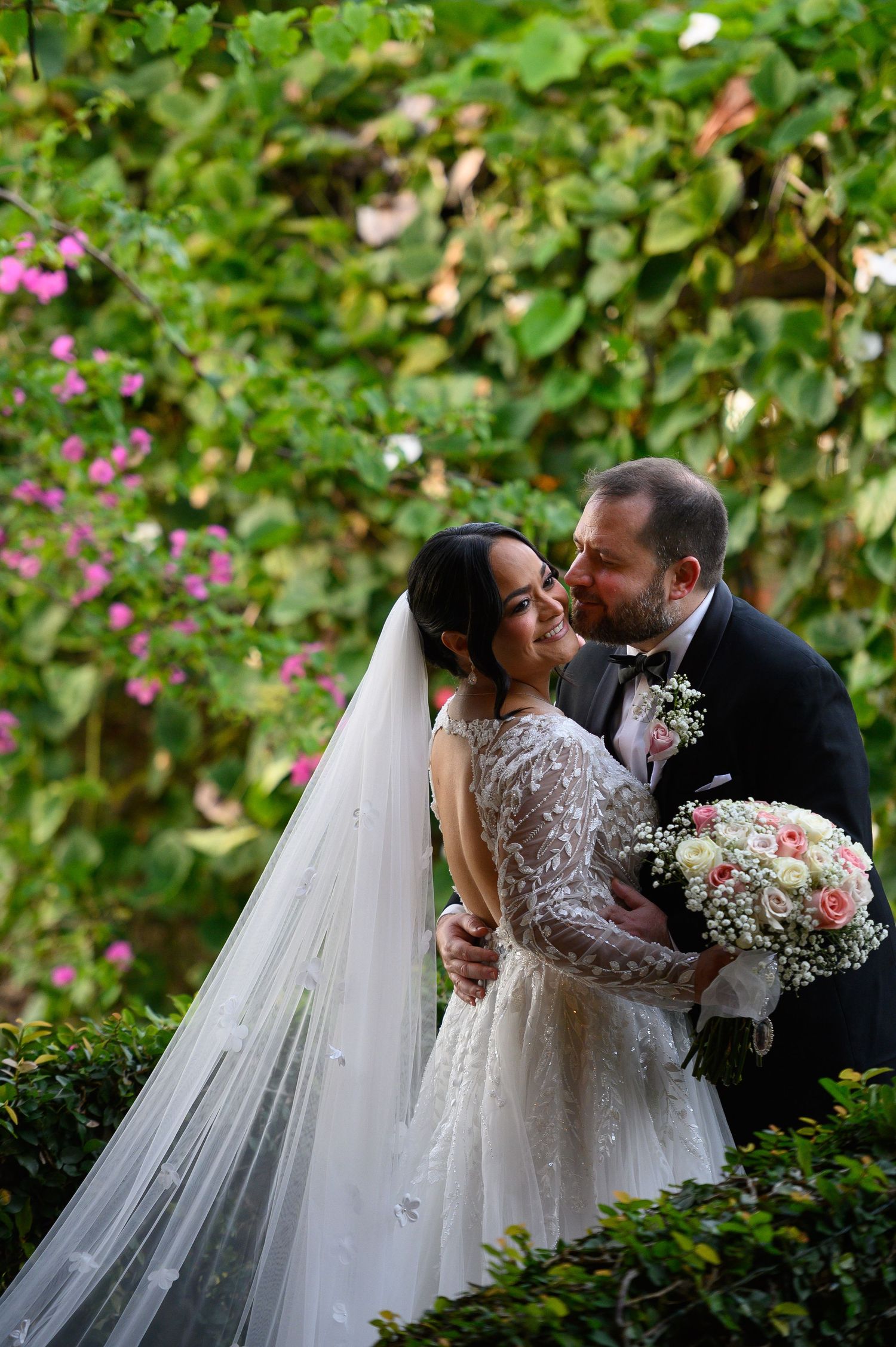 Groom kissing bride on cheek in lush garden setting, bride in beaded gown with veil holding pink and white bouquet.