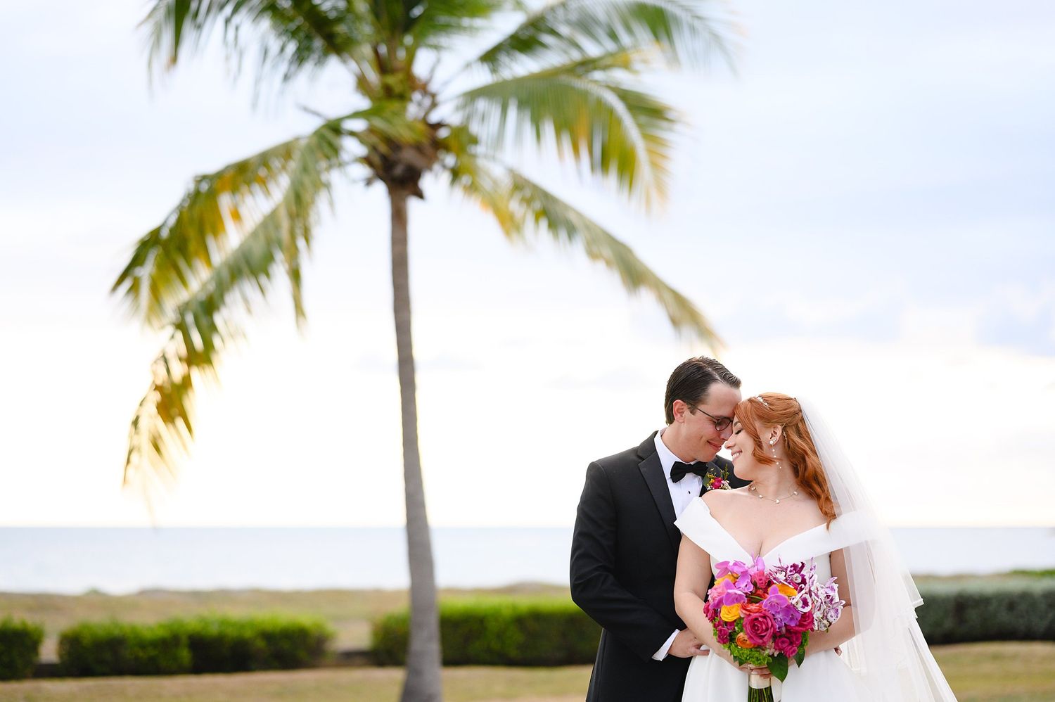 Bride and groom share a romantic kiss near a palm tree with ocean views on their tropical beach wedding day.