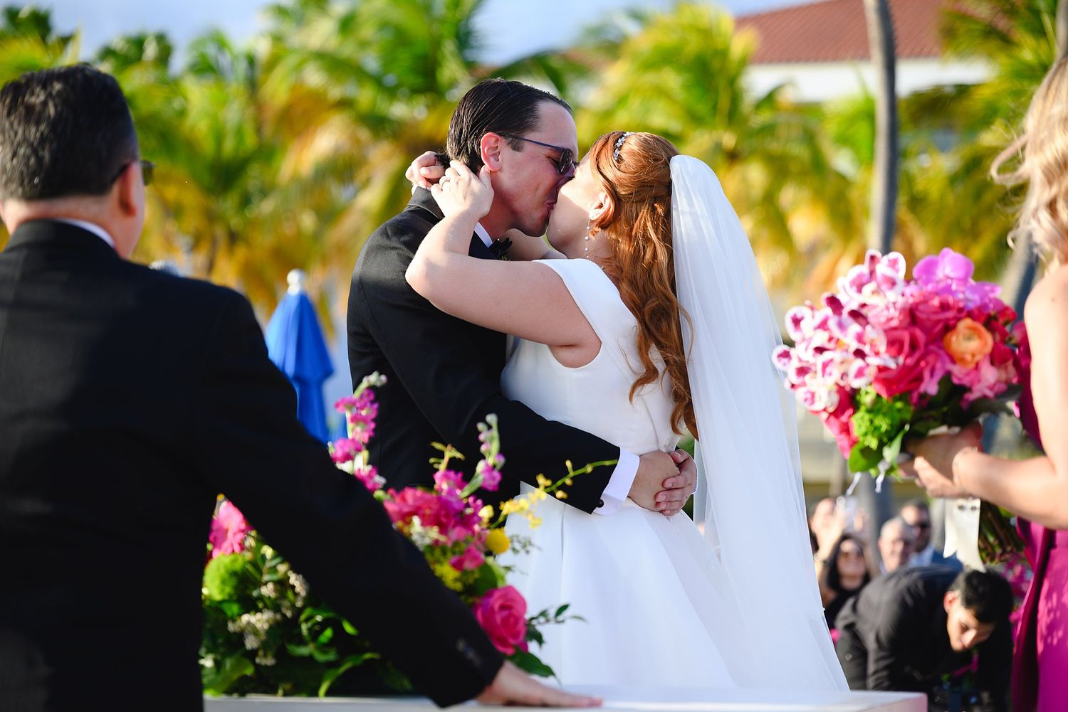 Bride and groom share first kiss at tropical outdoor wedding ceremony surrounded by vibrant pink floral arrangements.