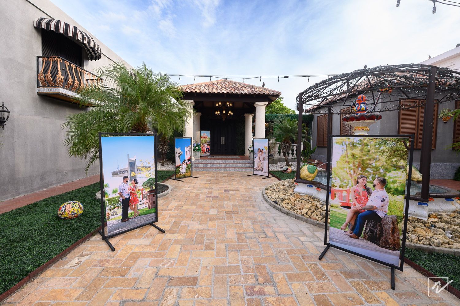Brick-paved courtyard with palm trees, framed family photo displays, and a covered pergola leading to a rustic stone entrance building.