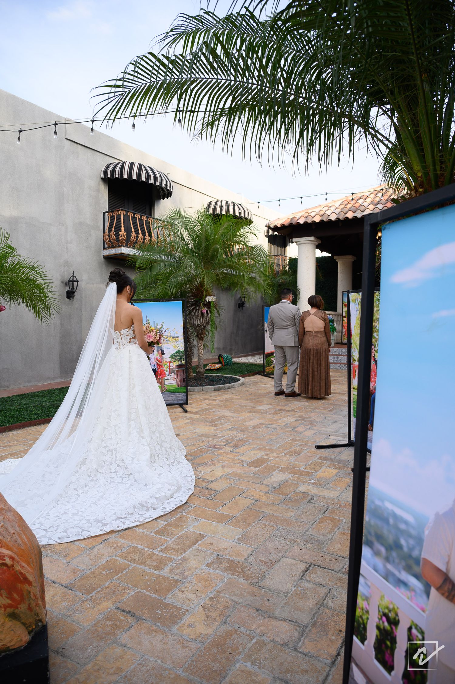 Bride in a long lace wedding gown with veil walks across a stone patio past framed photos toward guests under palm trees.