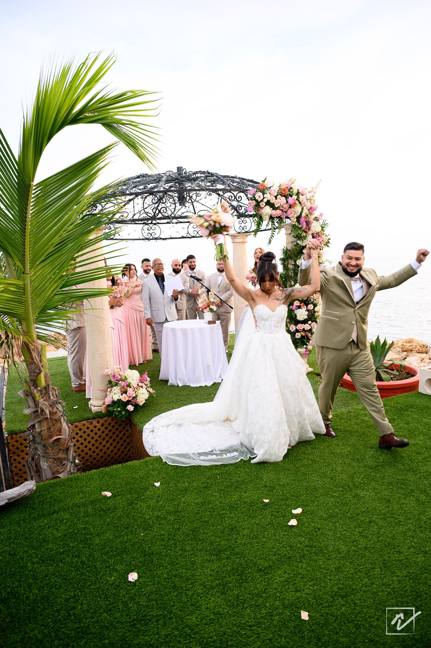 Bride in a flowing white gown and groom in a tan suit raise their arms in celebration after their oceanside wedding ceremony.