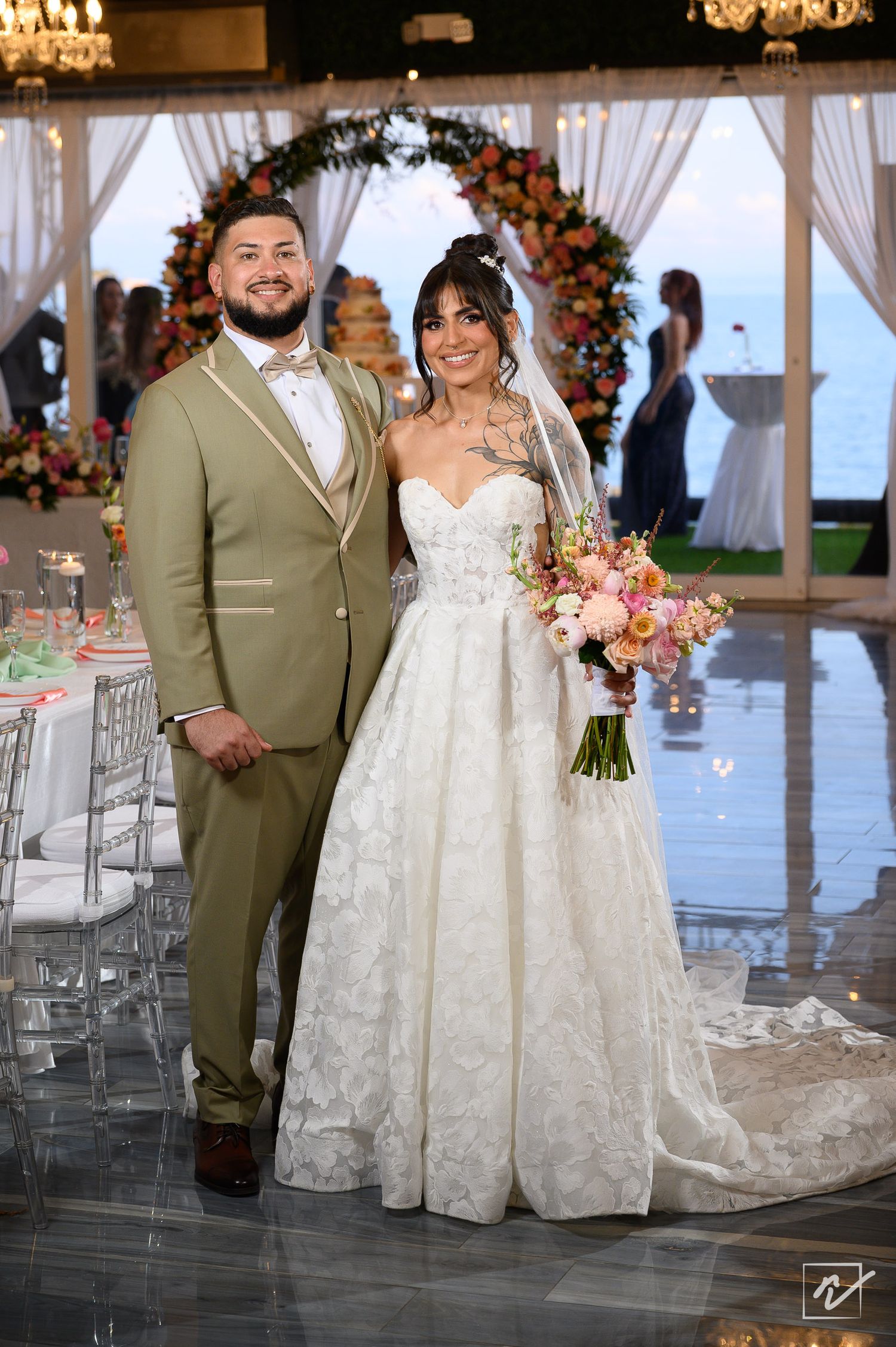 Smiling bride in a lace gown holding a pink bouquet stands beside a groom in an olive suit at a seaside wedding reception.