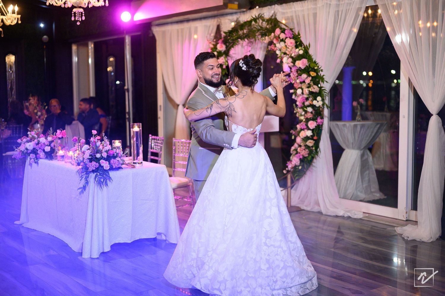 Bride in a white ball gown and groom share a first dance at an elegant wedding reception with floral arch and purple centerpieces.