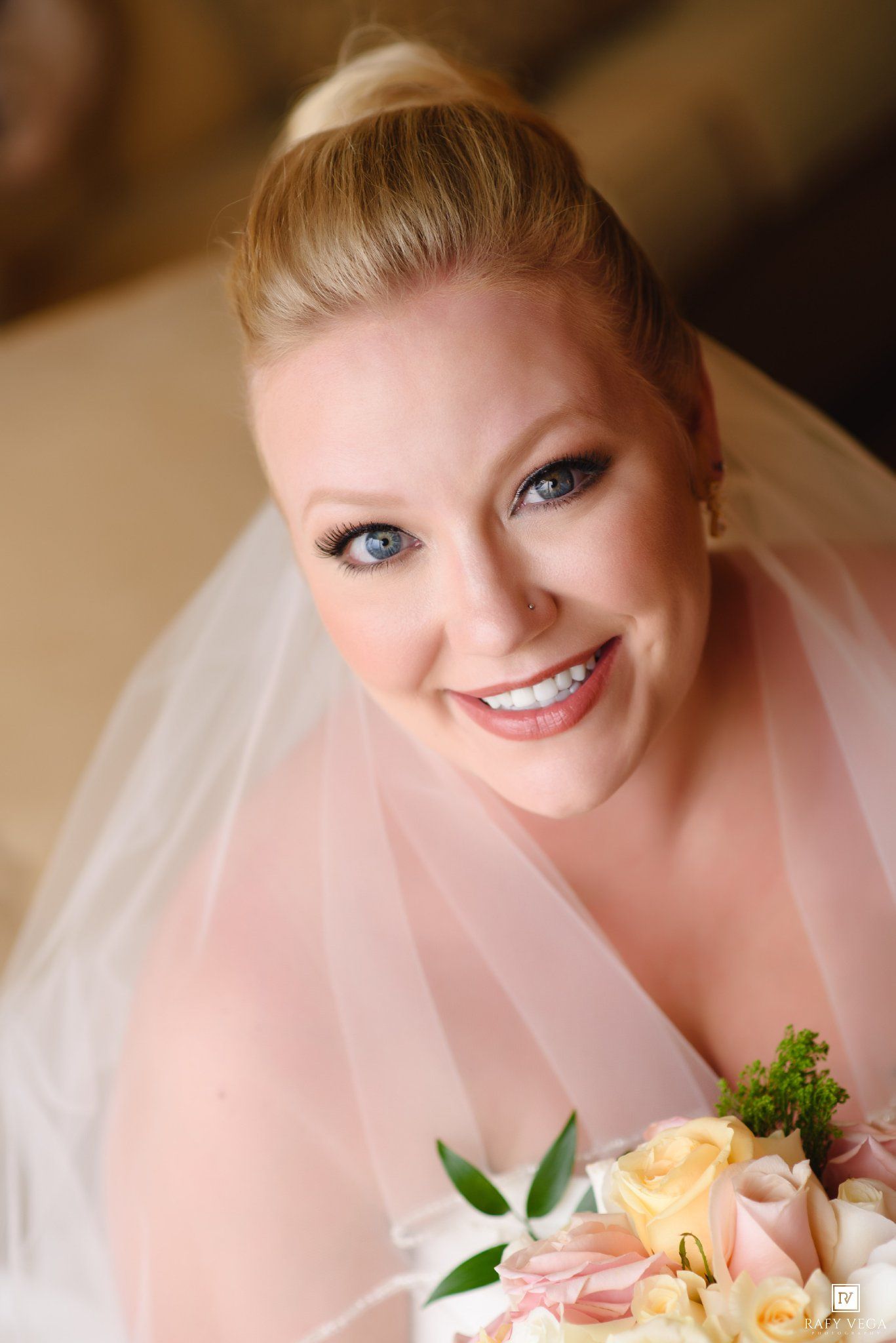 Smiling blonde bride with blue eyes wearing a white veil and holding a bouquet of pale pink and cream roses.