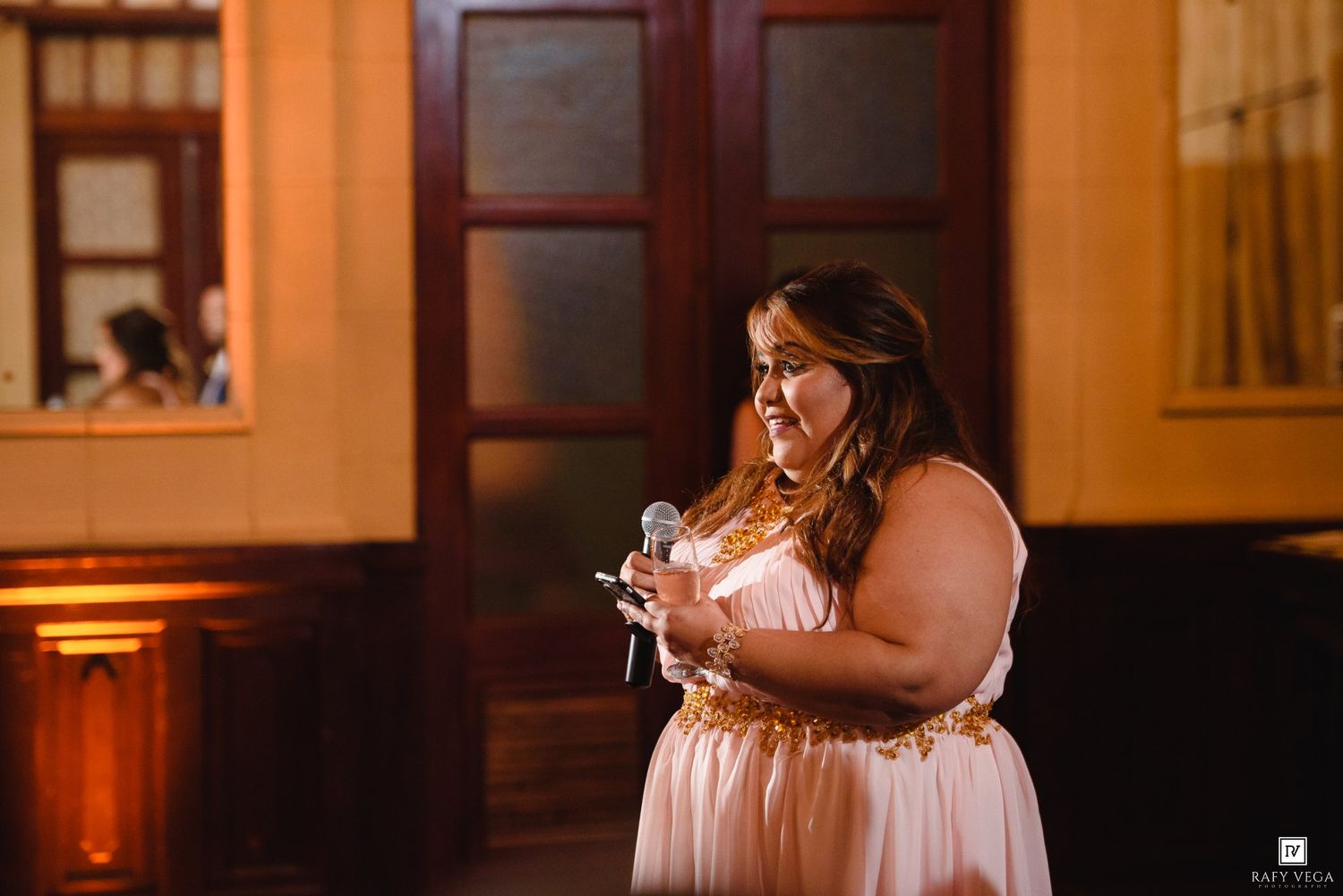 Woman in pink and gold dress speaks into microphone at elegant indoor event venue.