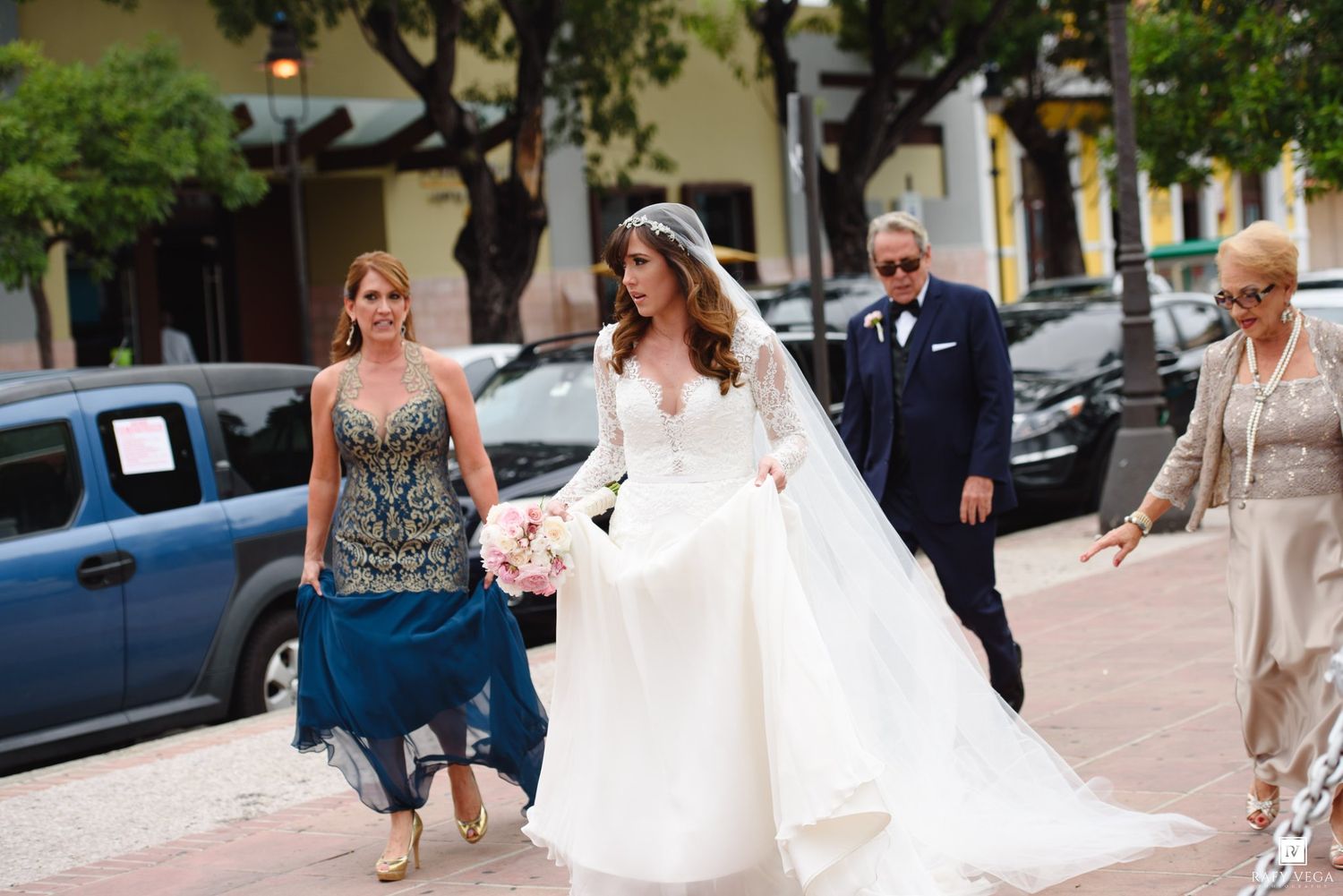 Bride in white gown with veil walks alongside family members in formal attire on a sunny outdoor street.