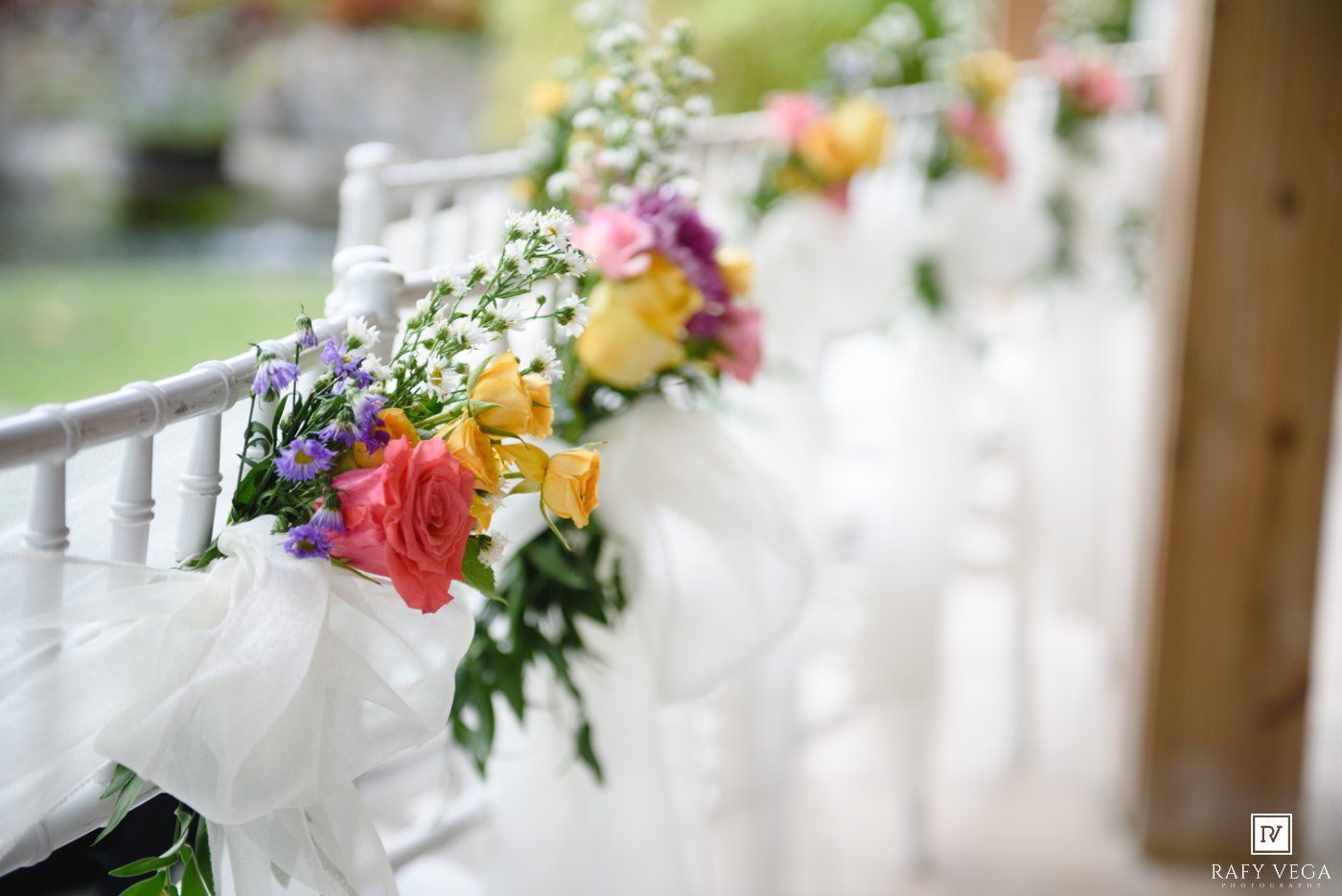 Colorful floral arrangements with roses and wildflowers tied with white tulle bows decorating white chairs at a wedding ceremony.
